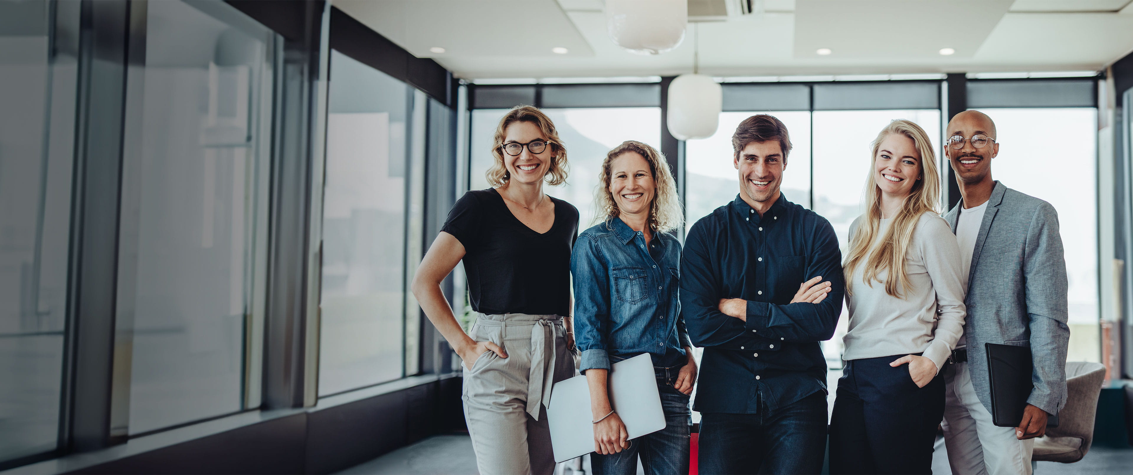 2N Cinq personnes d'affaires souriantes se tenant ensemble dans un espace de bureau moderne et lumineux avec de grandes fenêtres