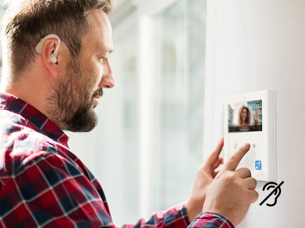 Man met een baard en een gehoorapparaat die een video-intercom gebruikt met iemand op het scherm en een symbool voor toegankelijkheid voor slechthorenden.
