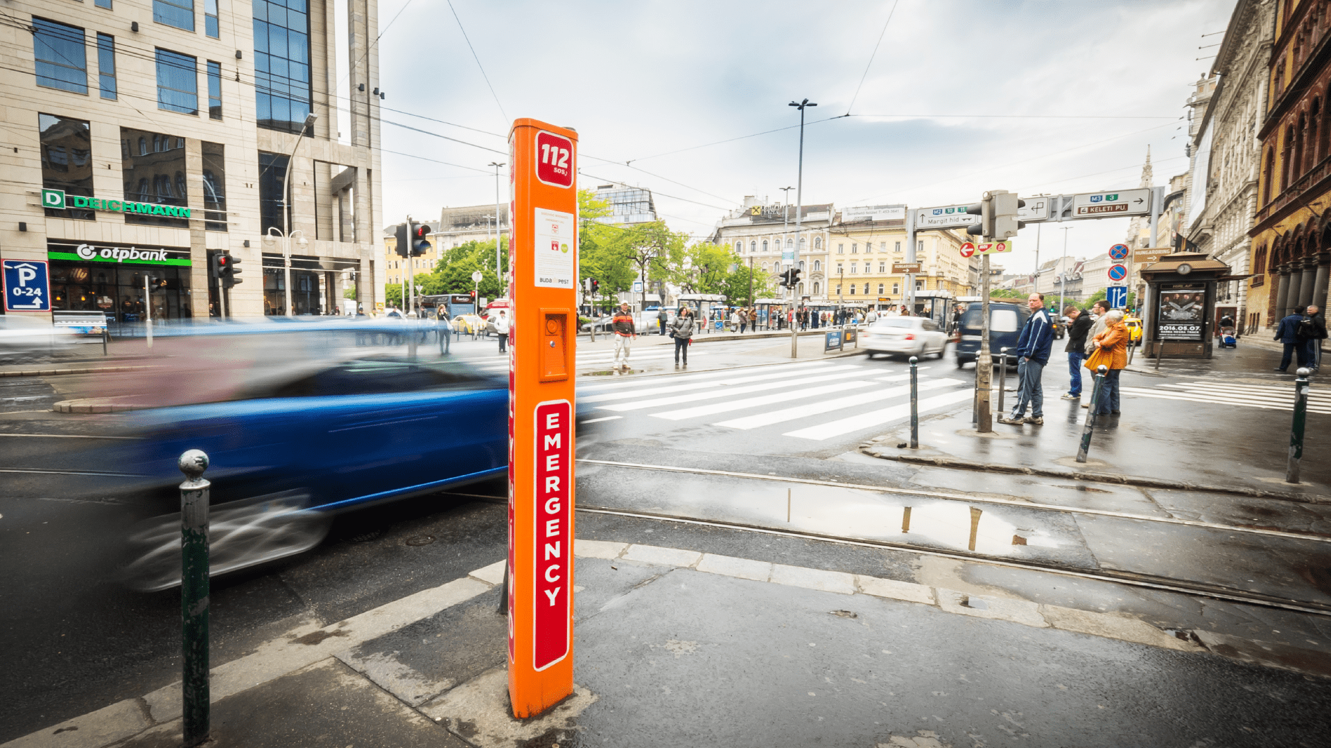 Une borne d'appel d'urgence orange est bien visible à un coin de rue très fréquenté, avec beaucoup de voitures et de piétons.