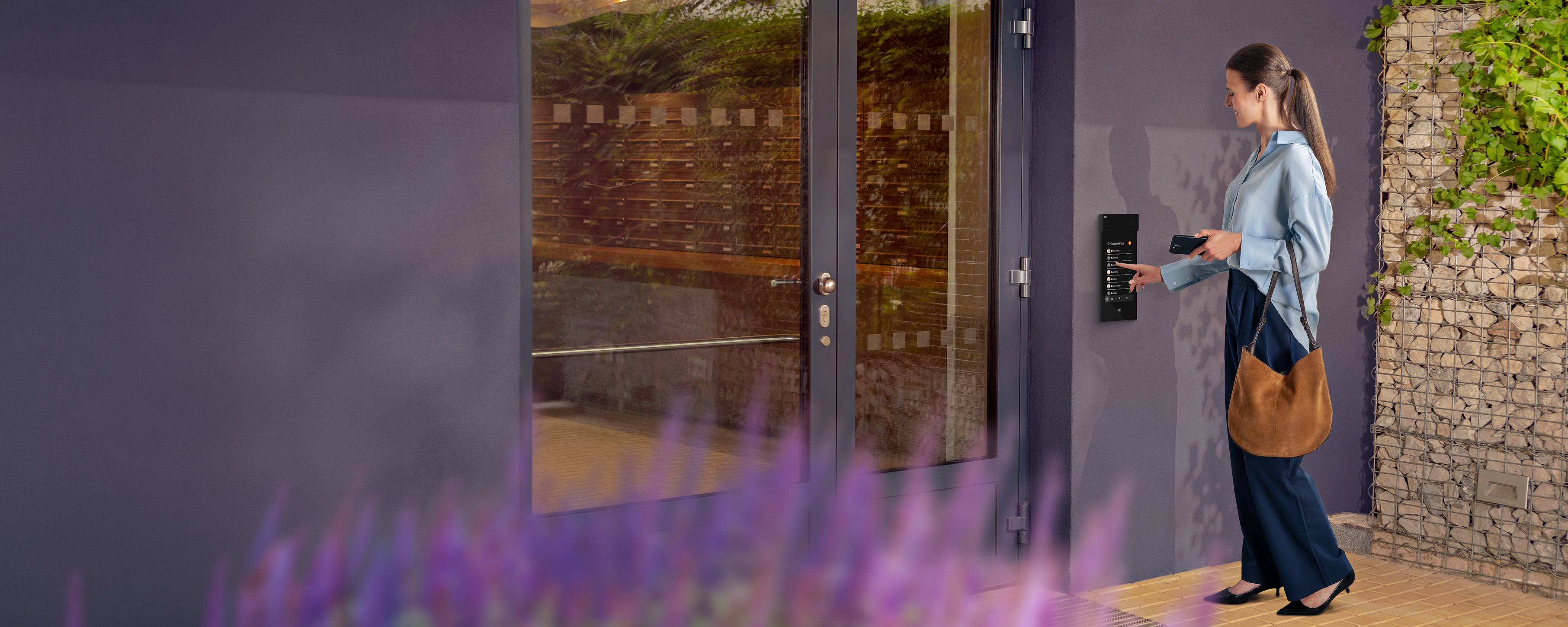 Person using a smartphone and key fob to access a modern glass entrance door with a wall-mounted reader.