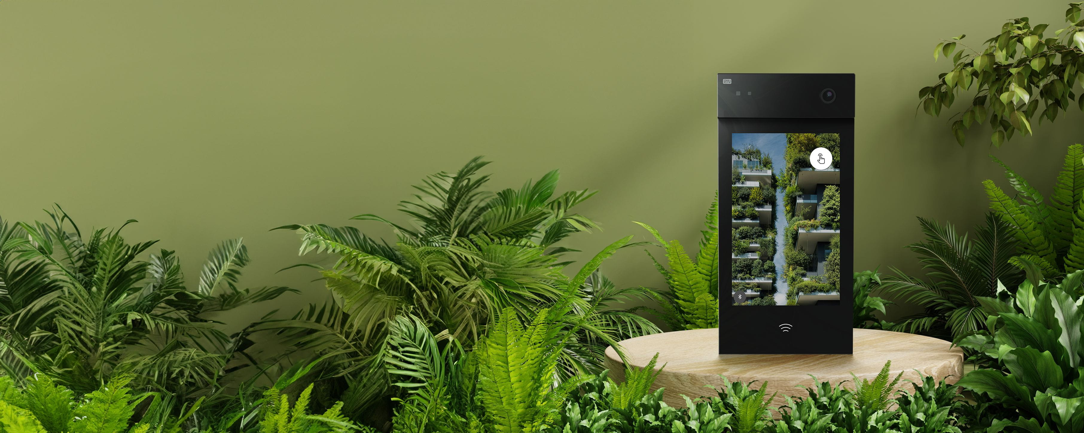 Black 2N intercom with a display shown on a circular wooden pedestal surrounded by lush green plants.