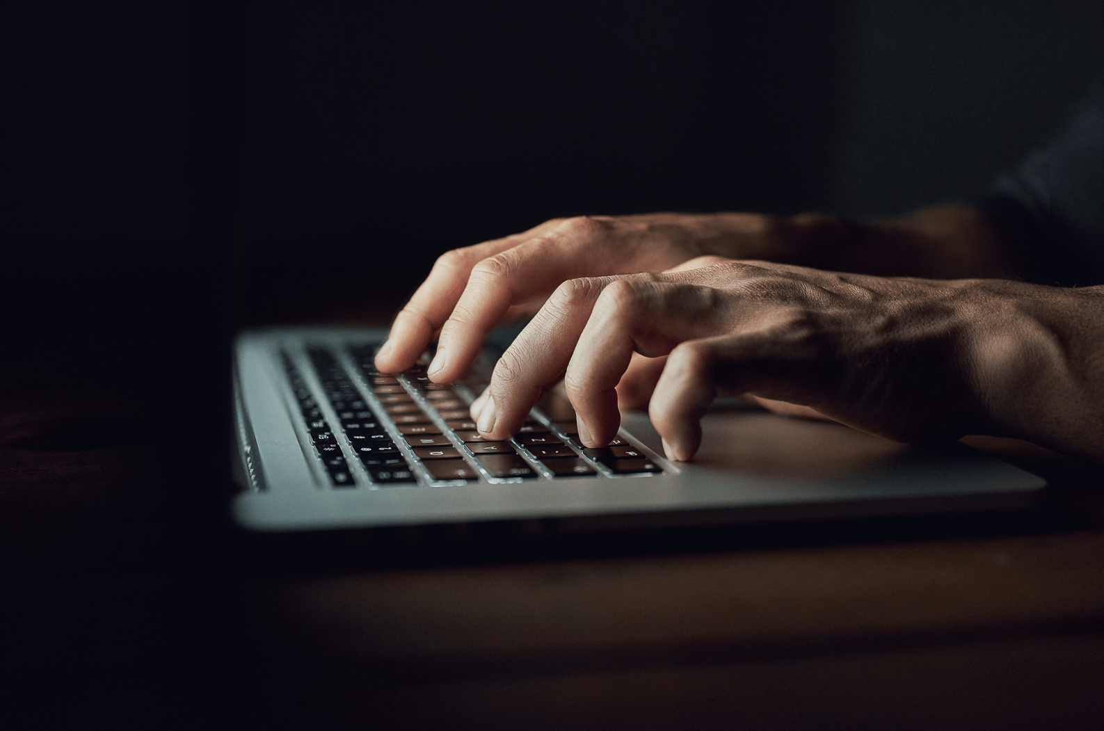 Hände, die auf einer Laptop-Tastatur in einem schwach beleuchteten Arbeitsbereich tippen.