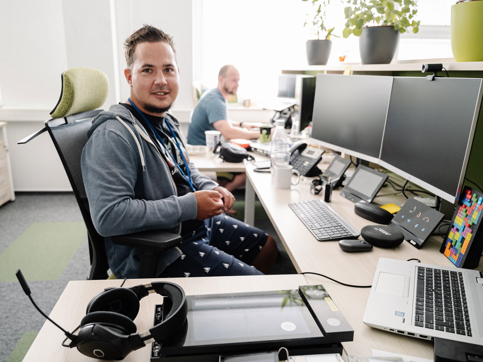 Ein lächelnder Typ in Shorts und Kapuzenpulli sitzt an einem Arbeitsplatz mit mehreren Monitoren in einem hellen Büro.