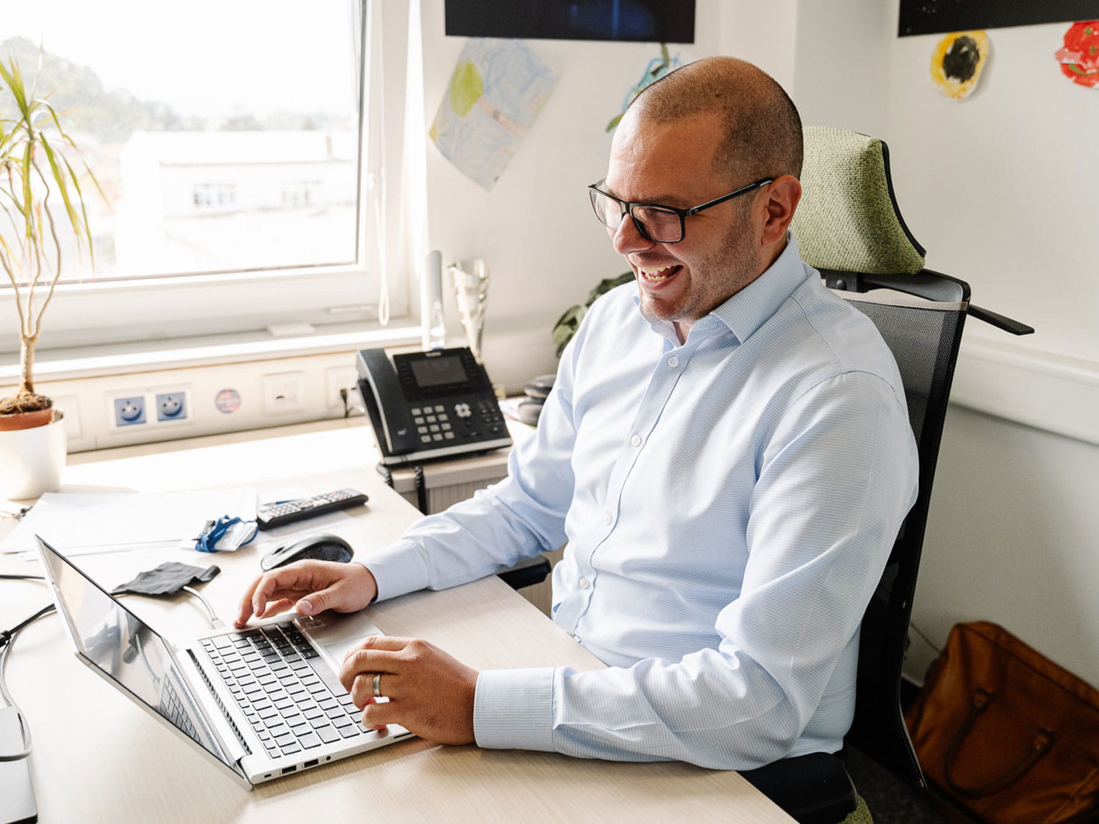 Ein Mann mit Glatze, Brille und blauem Hemd lächelt, während er an seinem Schreibtisch am Laptop arbeitet.