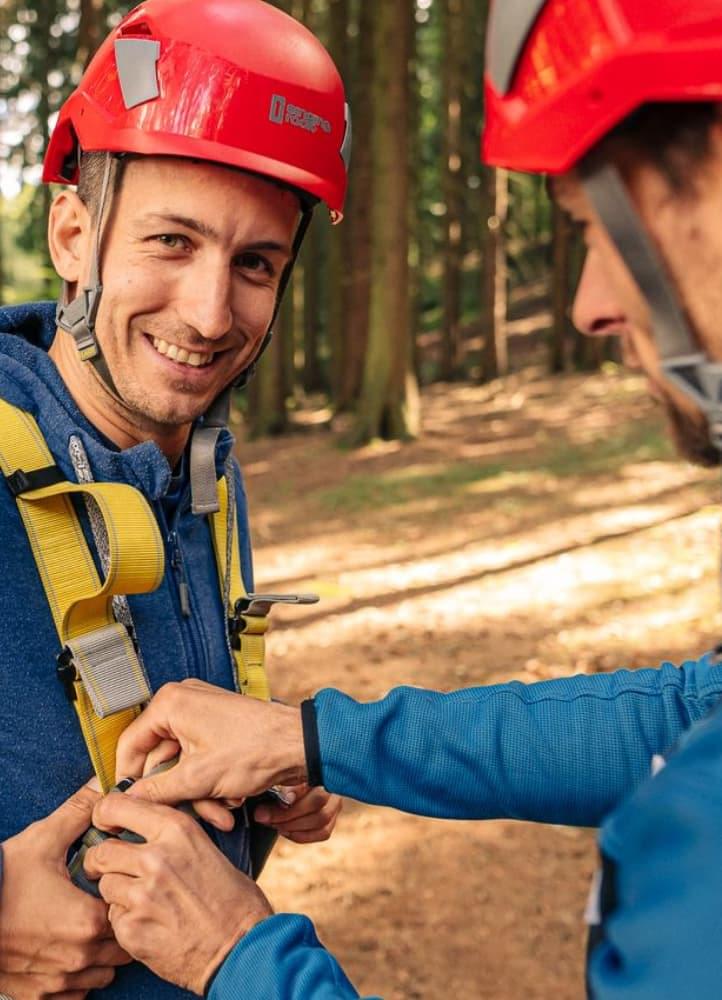 Ein lächelnder Mann in einem roten Helm lässt seinen Sicherheitsgurt von einem Ausbilder im Wald anpassen.