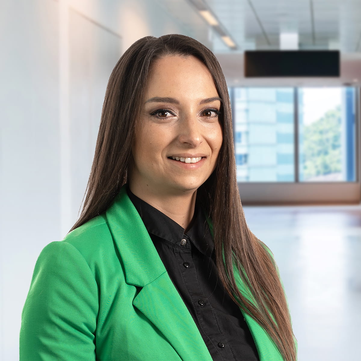 Porträt einer Person in einem grünen Blazer, die in einem modernen Büro mit Glaswänden und sanfter Beleuchtung steht.