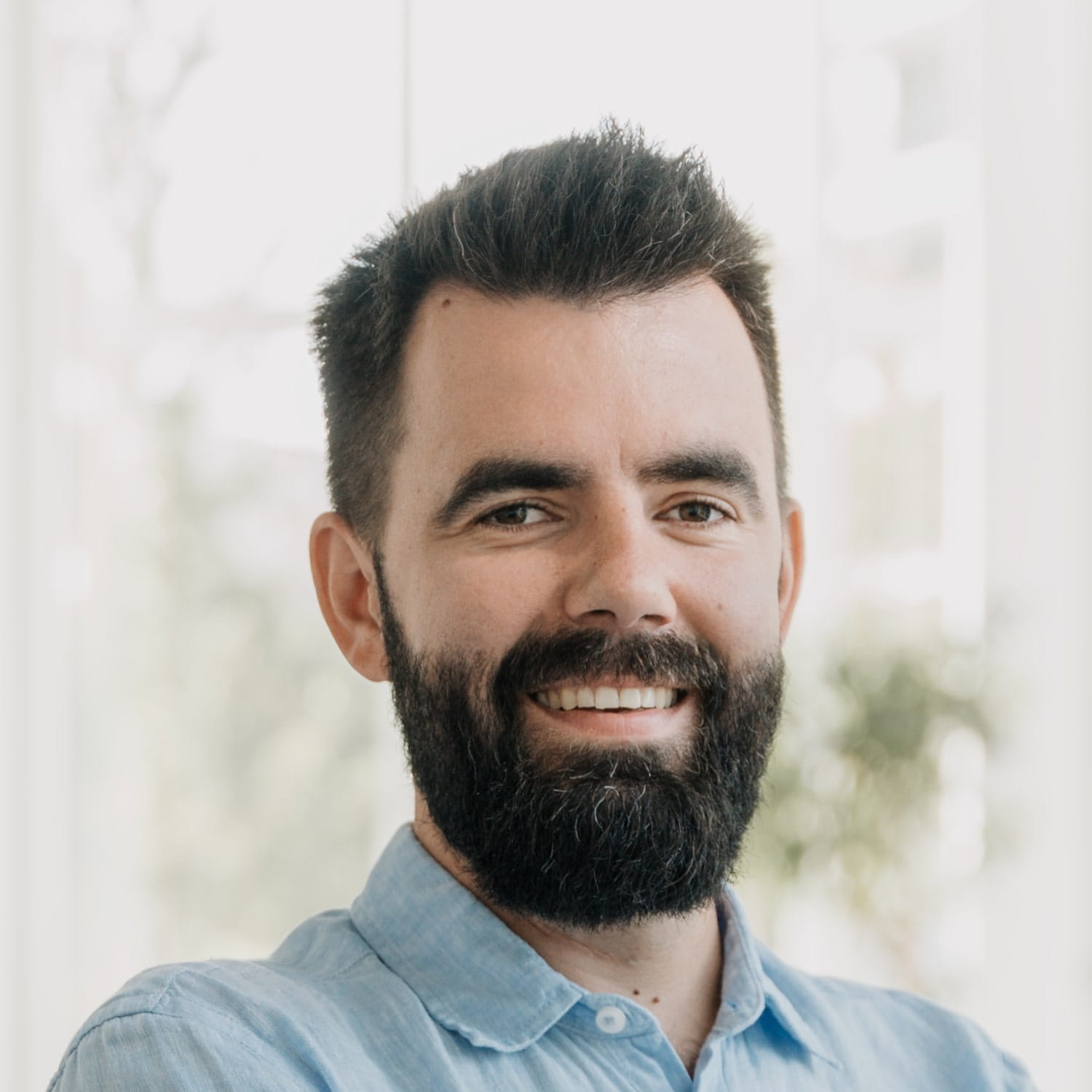 Photo d'un homme souriant avec une barbe fournie et des cheveux foncés portant une chemise à col bleu clair.