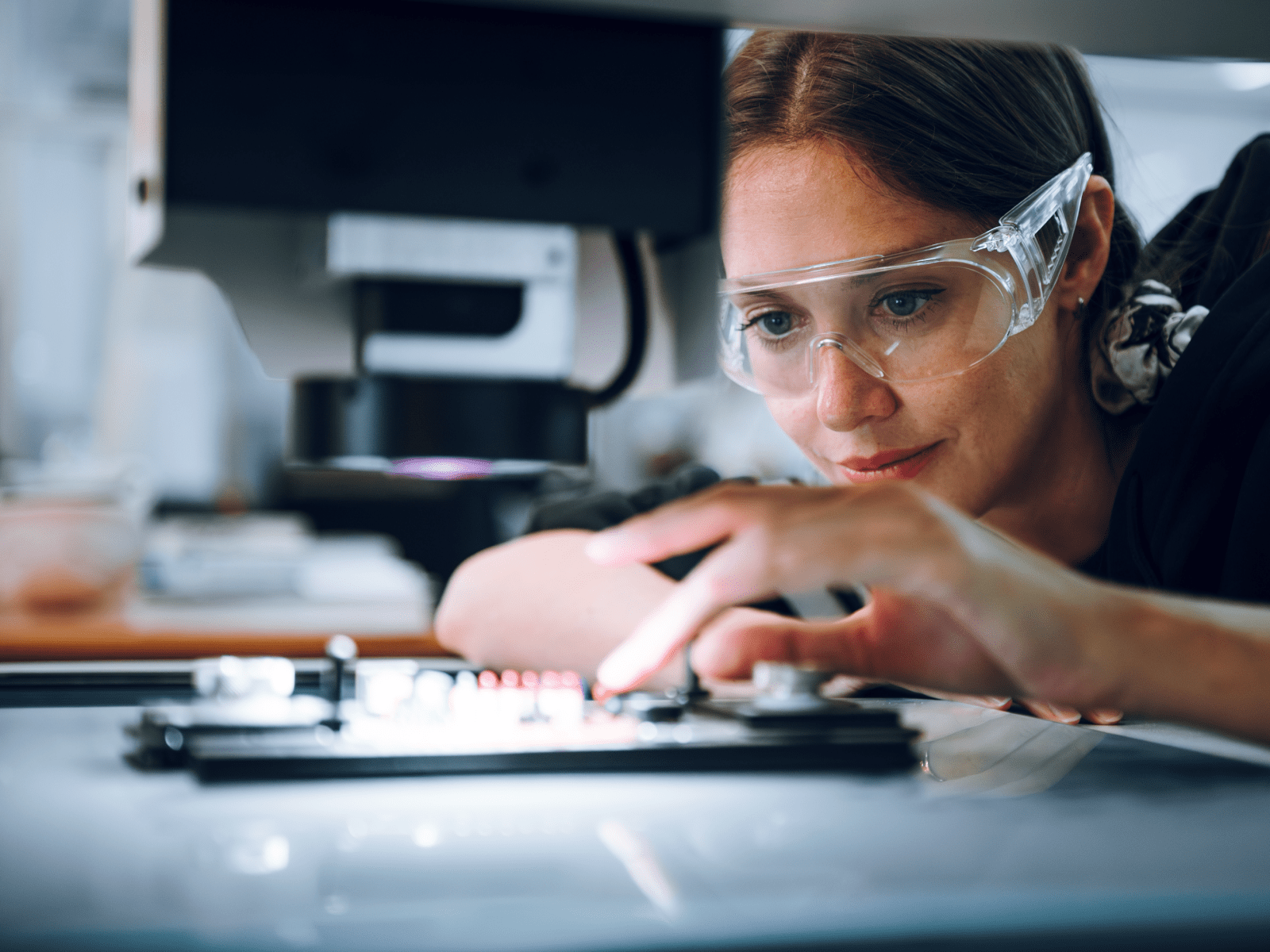 Une ingénieure avec des lunettes de sécurité qui bosse sur un circuit imprimé sous un éclairage industriel.