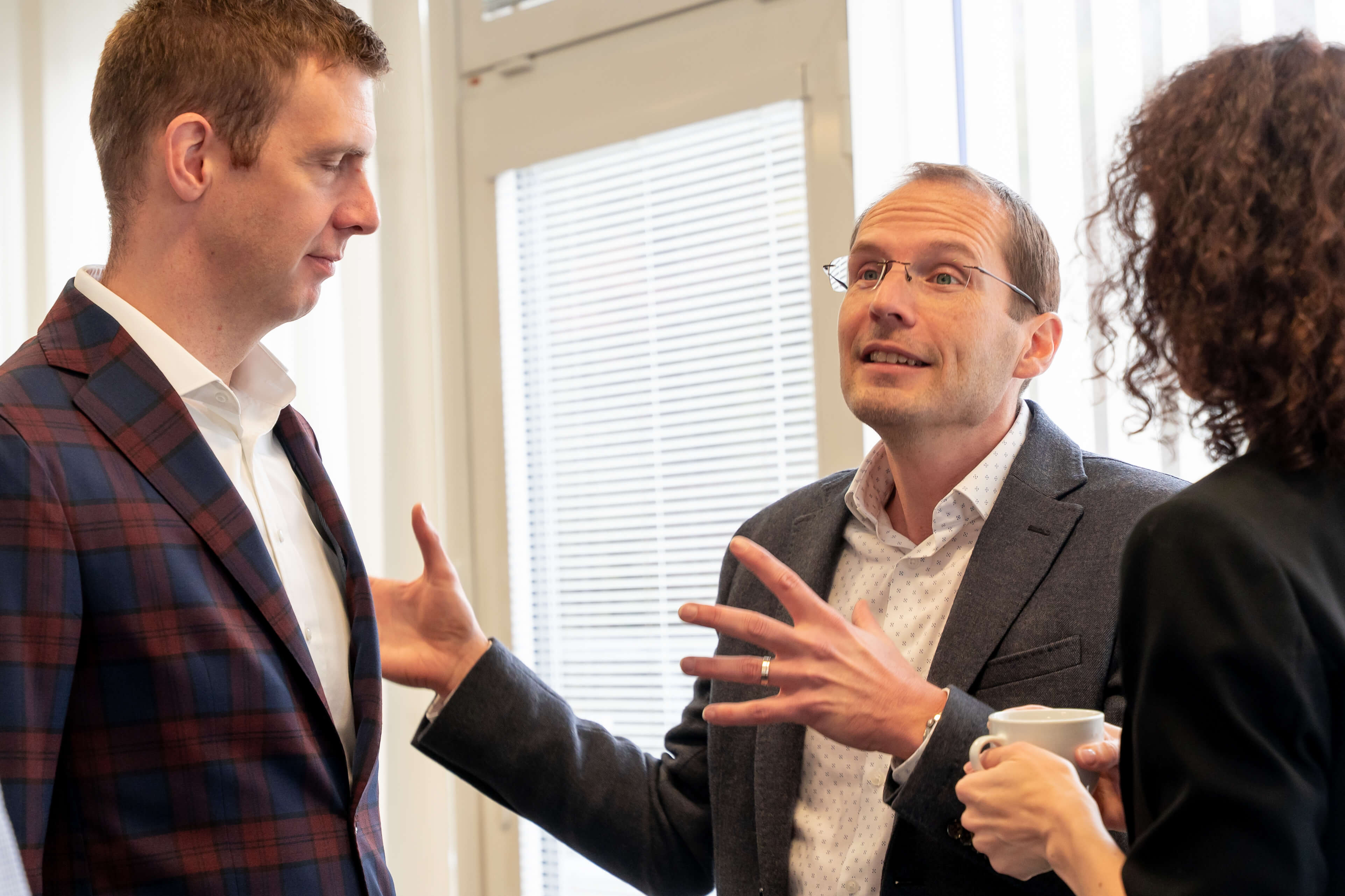 Three business people engaged in a discussion; one man gestures while holding a coffee cup.