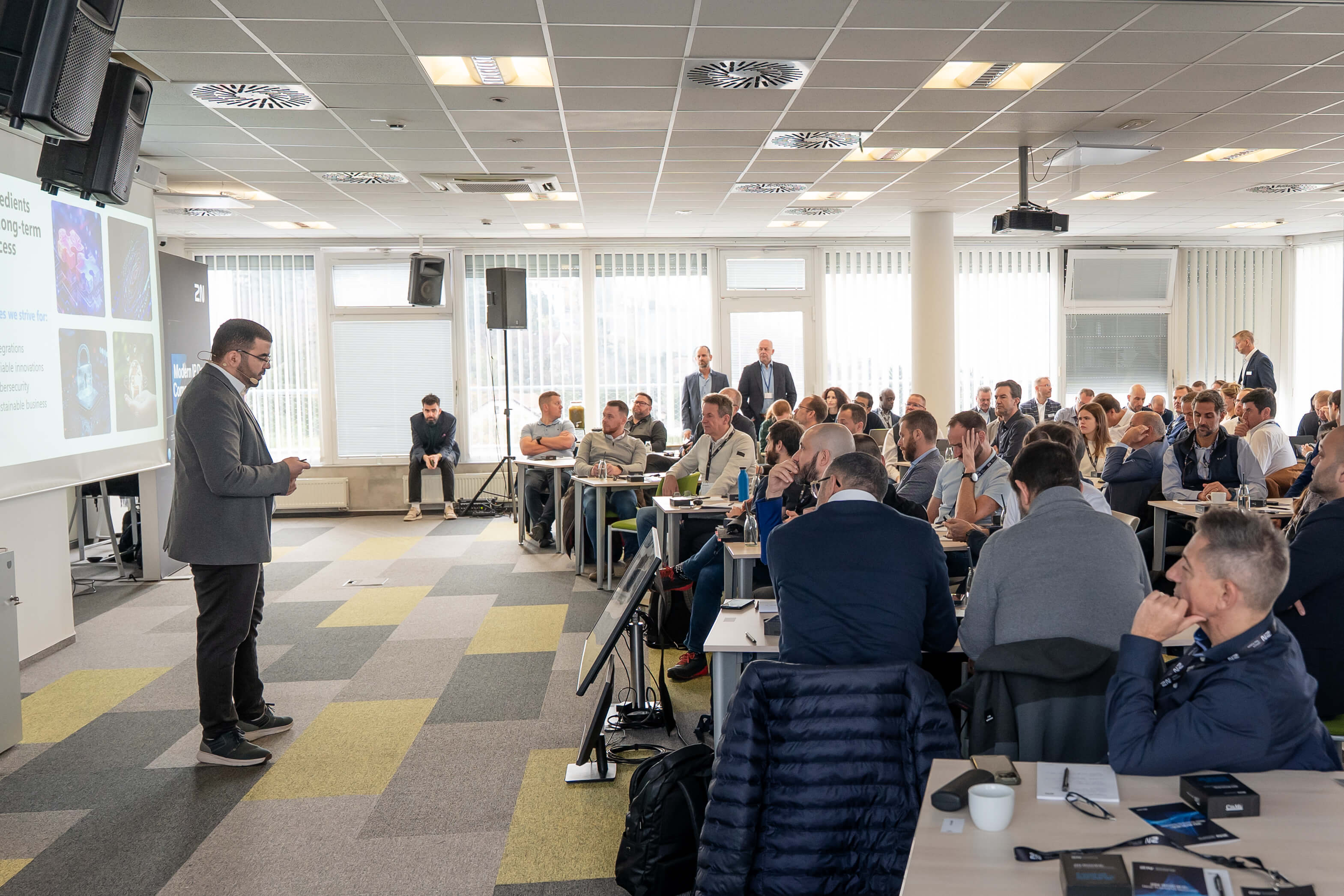 A speaker presents to a large audience seated at tables during a professional conference or seminar.