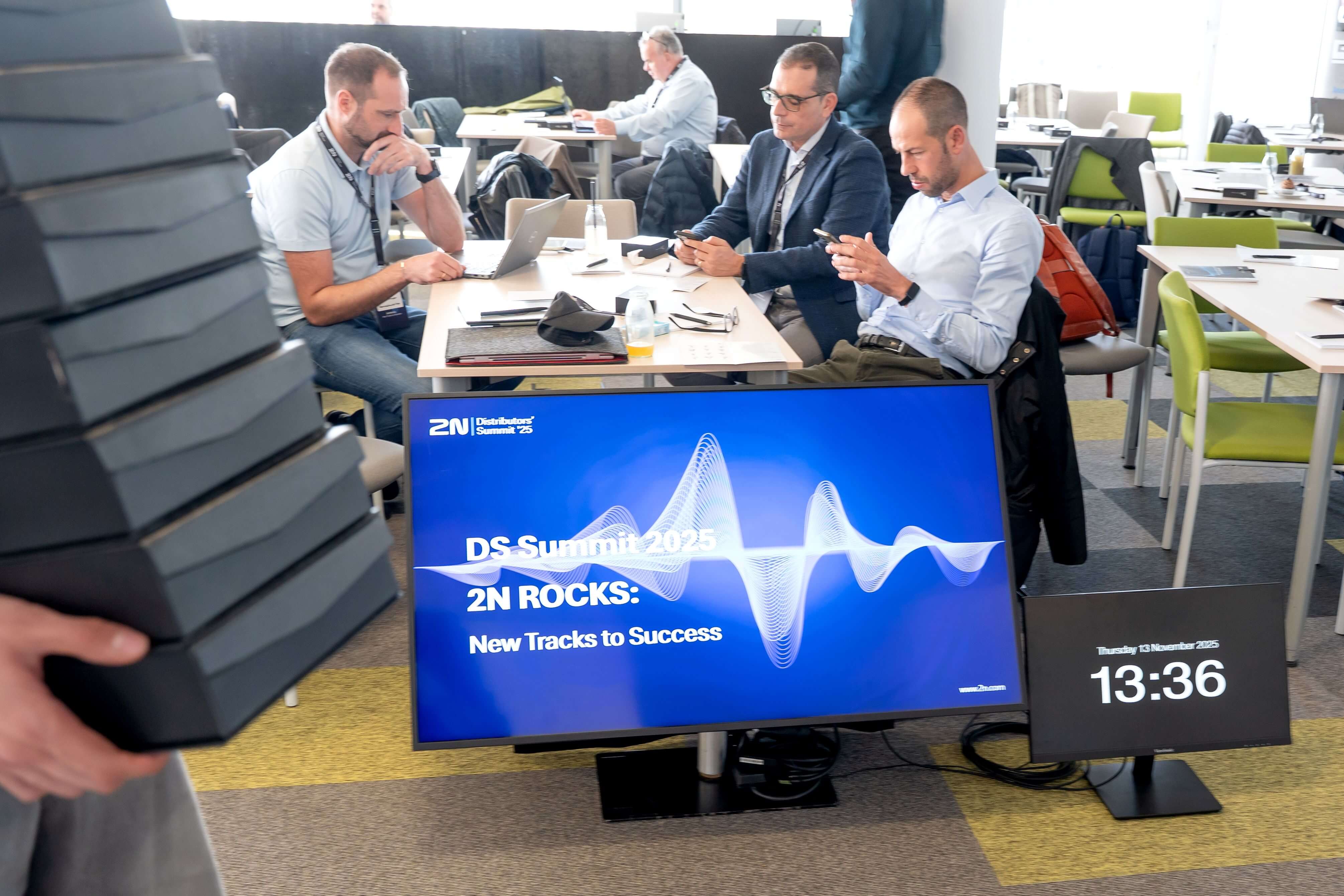 People sitting at a table with a screen showing "2N ROCKS: New Tracks to Success" and a digital clock.