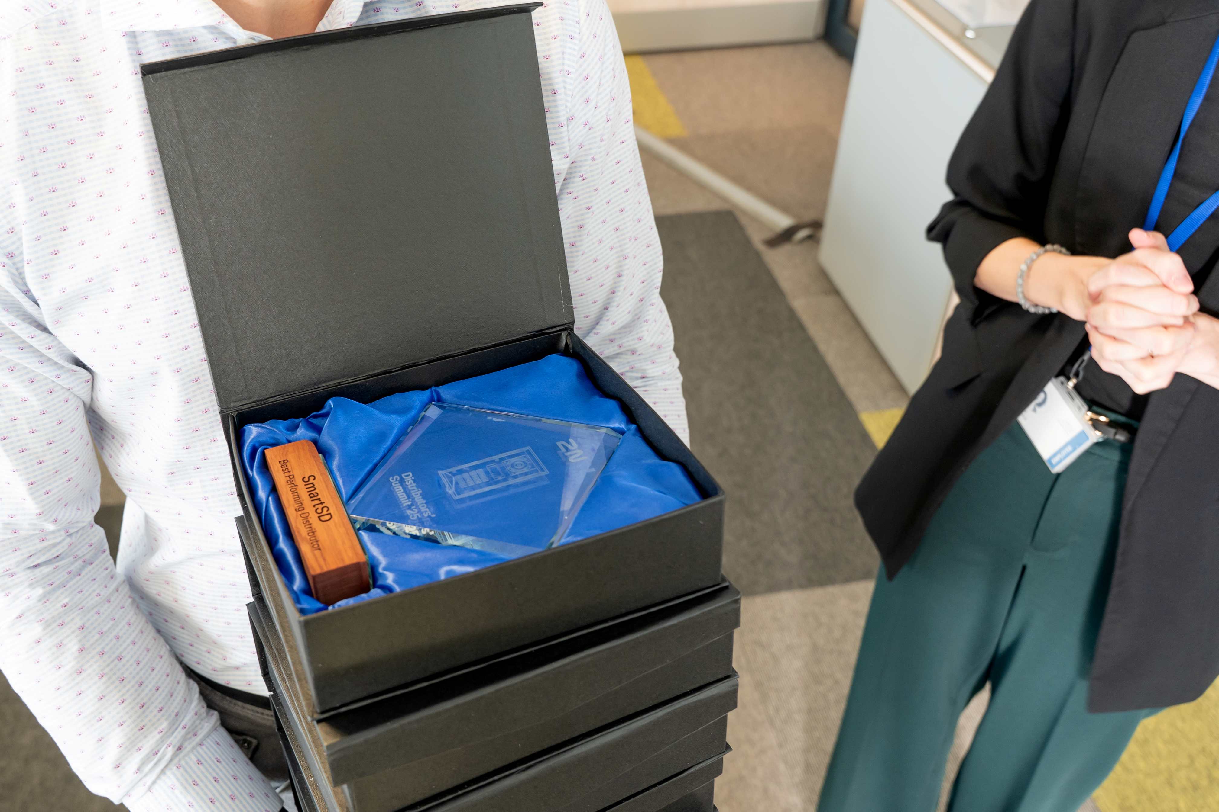 Close-up of a person holding a stack of black presentation boxes, with one open revealing a glass award and a wooden object on blue fabric.