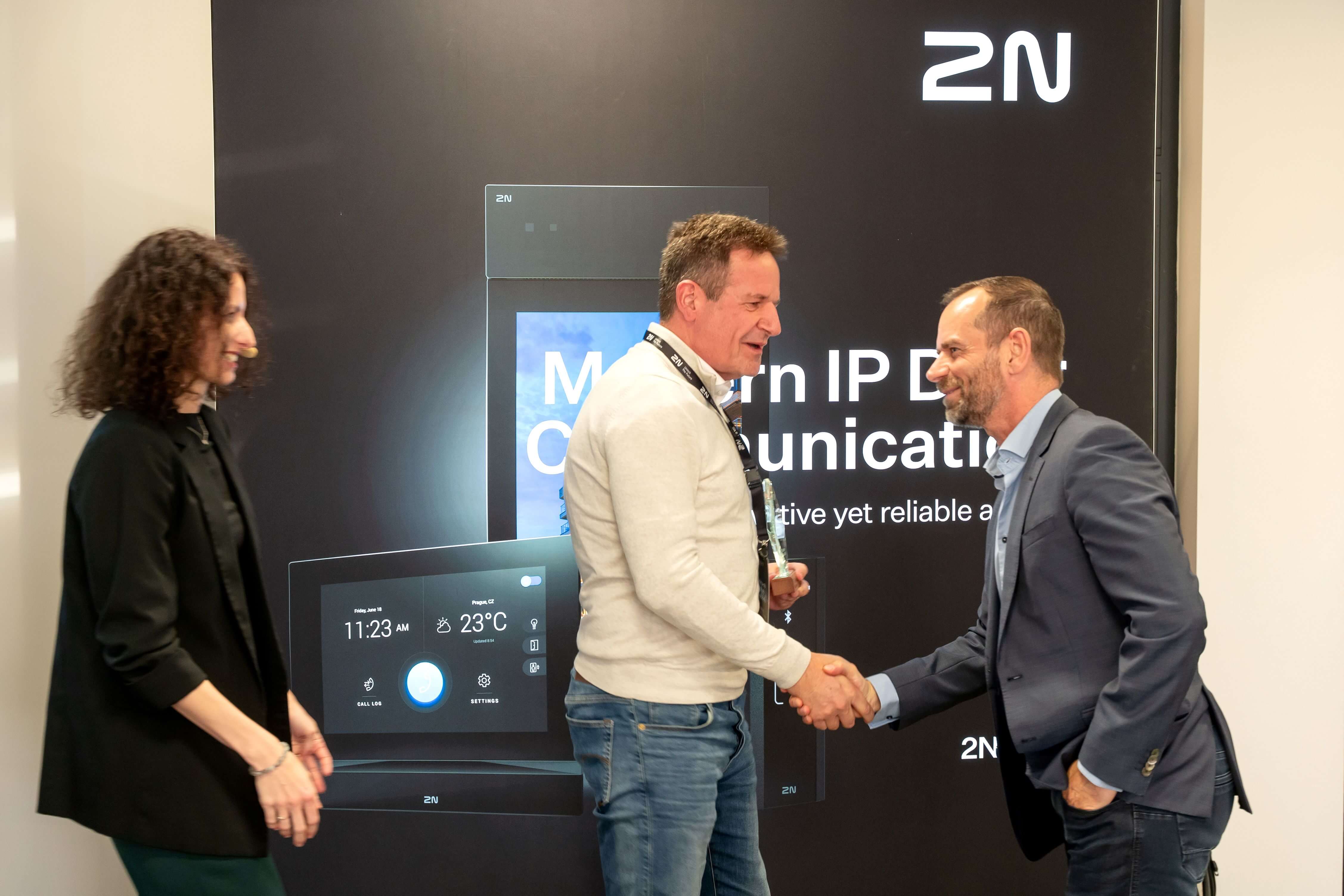 Two men shaking hands in front of a 2N "Modern IP Door Communication" display banner, with a woman beside them.