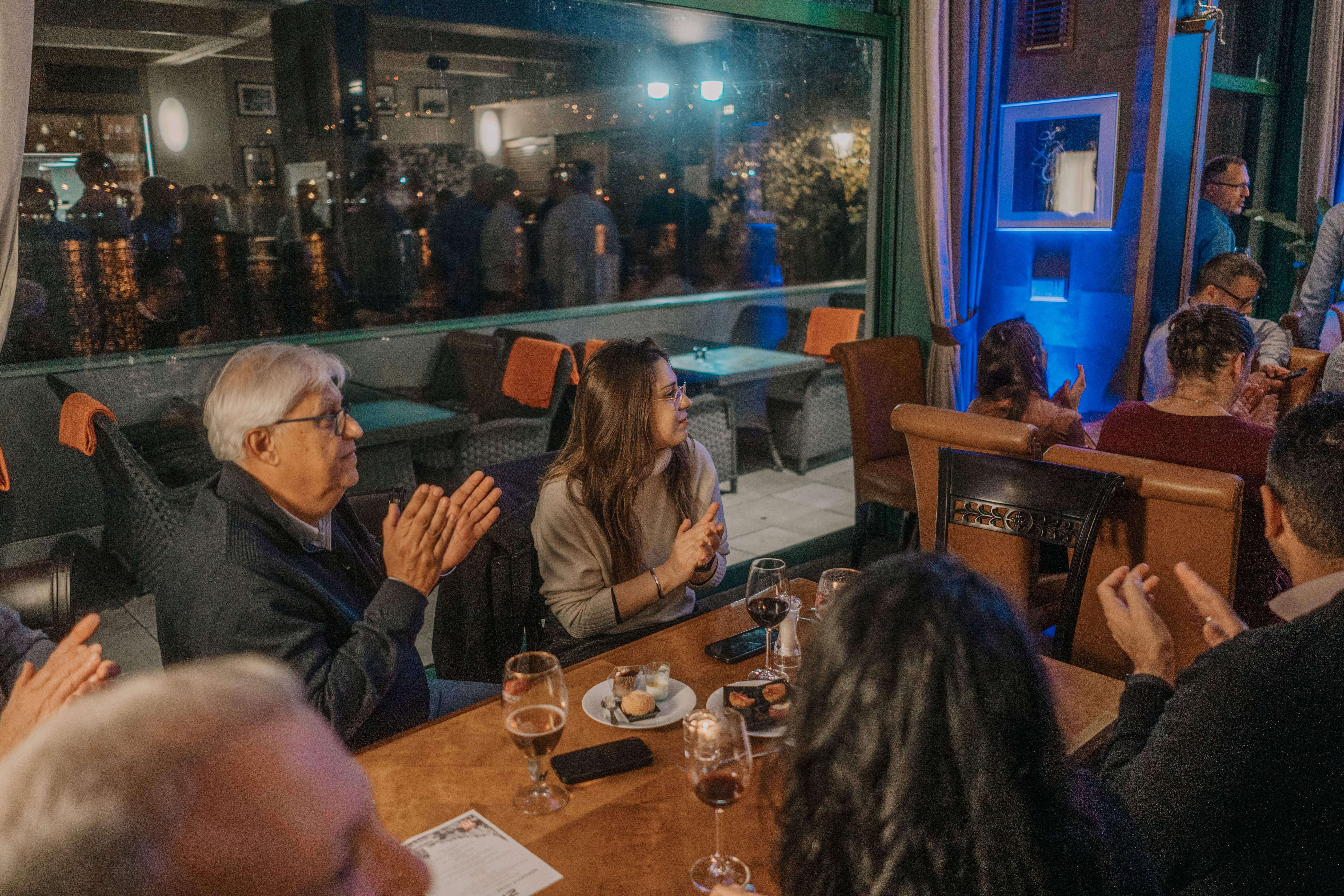 Guests at a dinner party sitting at tables, clapping and watching a performance in a dimly lit restaurant.