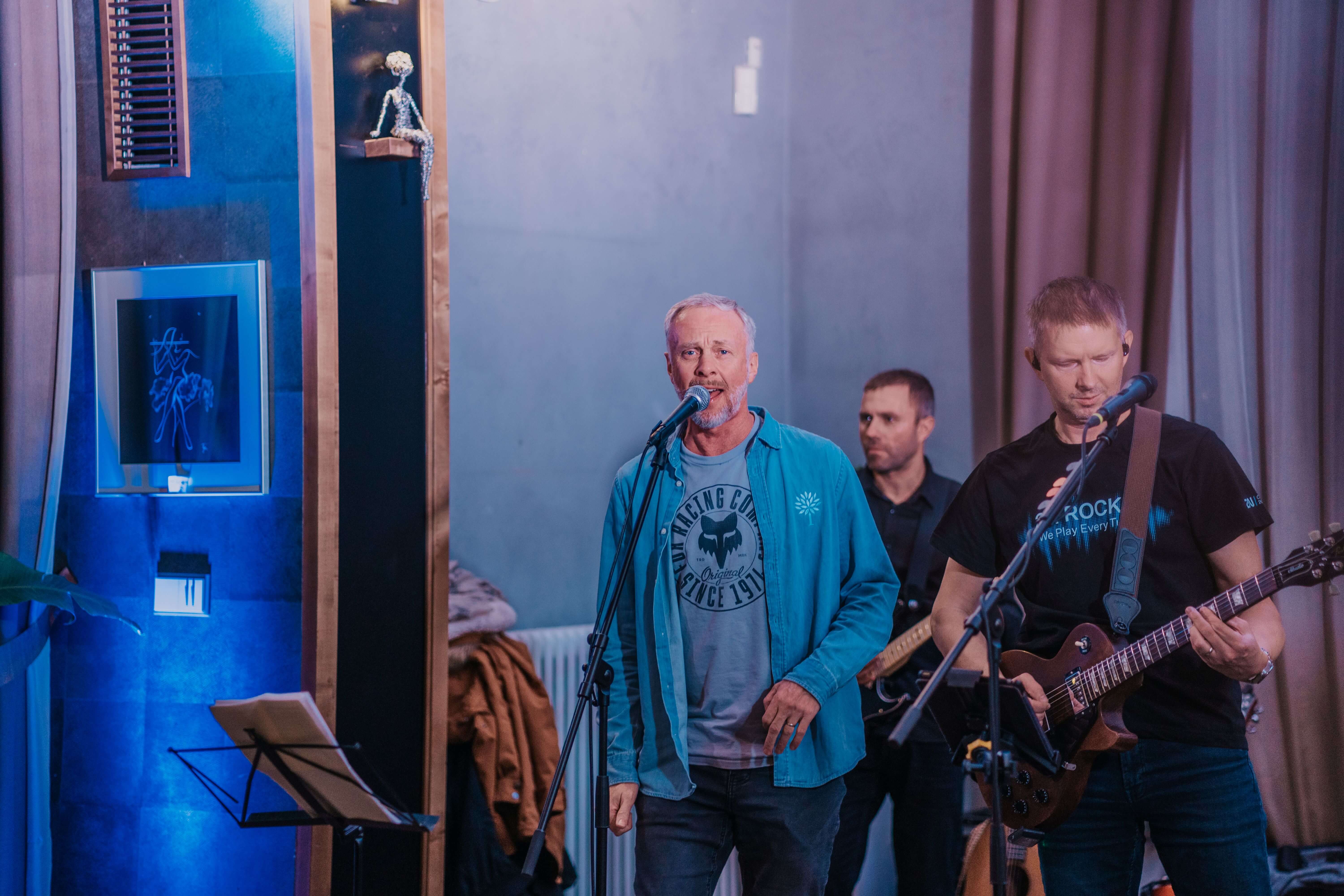 The lead singer in a blue shirt and grey t-shirt performs on stage with a guitarist and another band member in blue light.