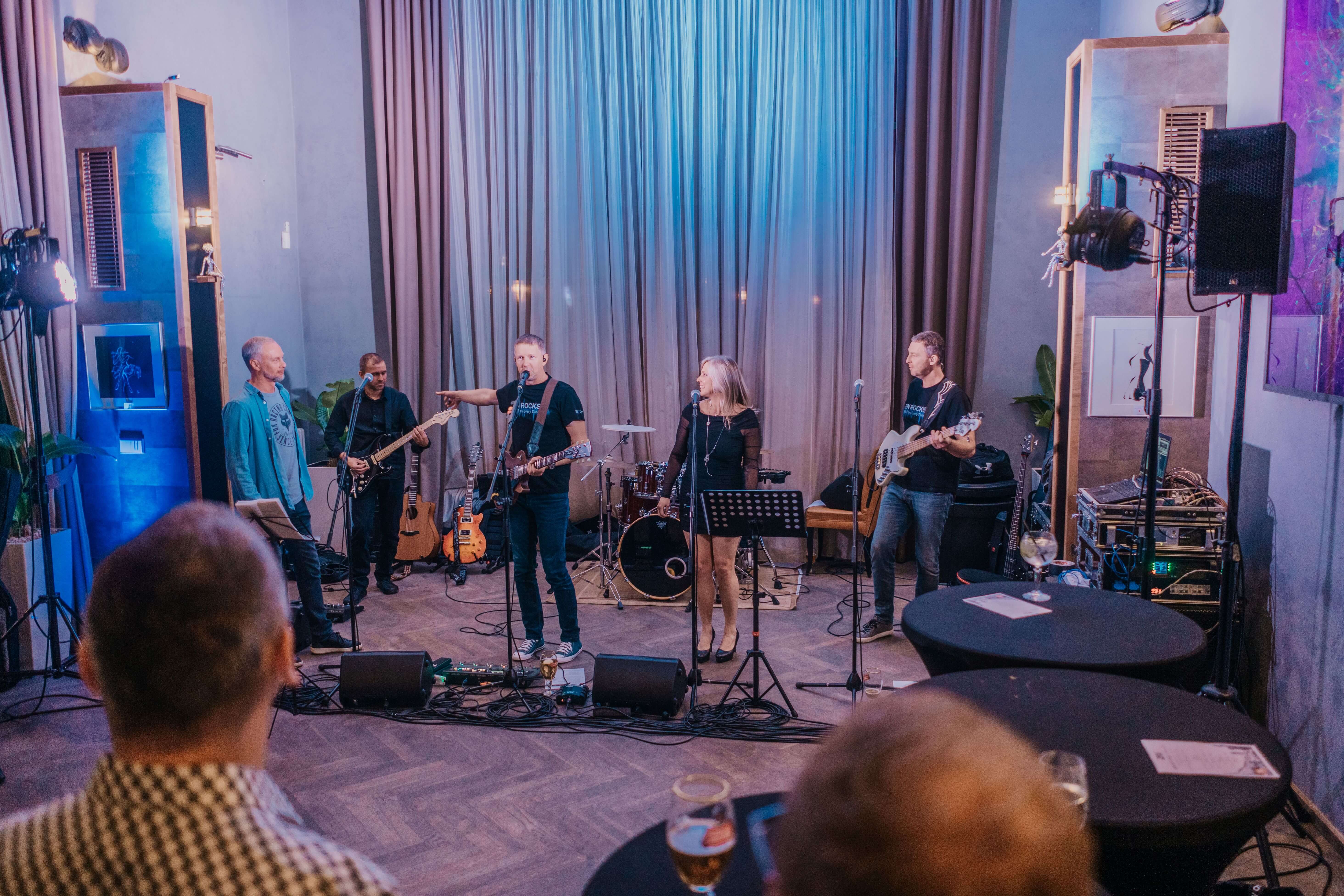 A five-person band performs on a stage with blue uplighting and curtains for an audience seated in the foreground.