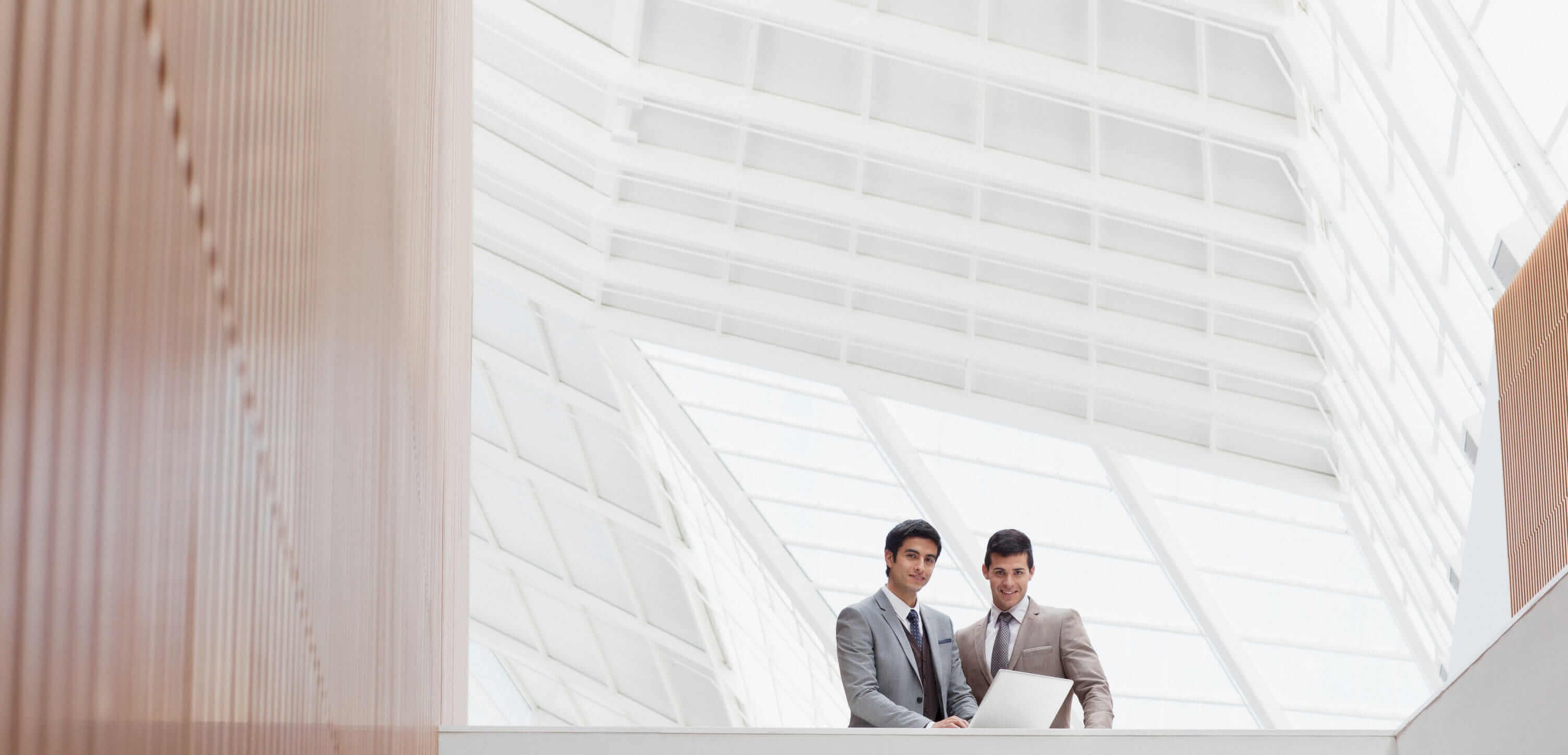 Deux gars en costume regardent un ordi portable dans l'atrium d'un immeuble de bureaux moderne et lumineux avec des boiseries.