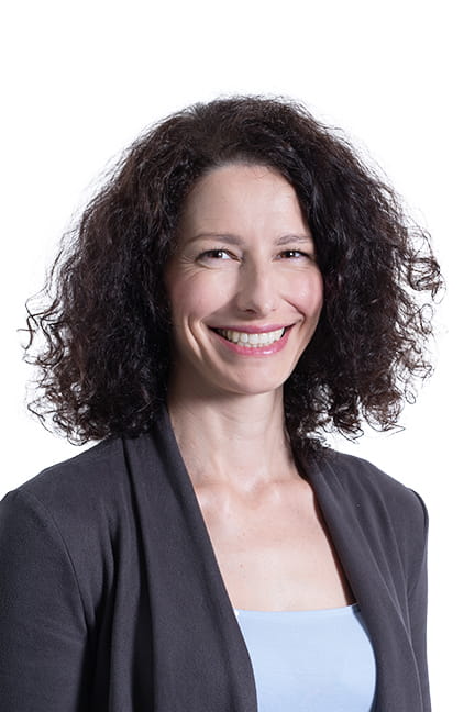 Photo pro d'une femme souriante aux cheveux foncés et bouclés, avec un haut bleu et un cardigan foncé.