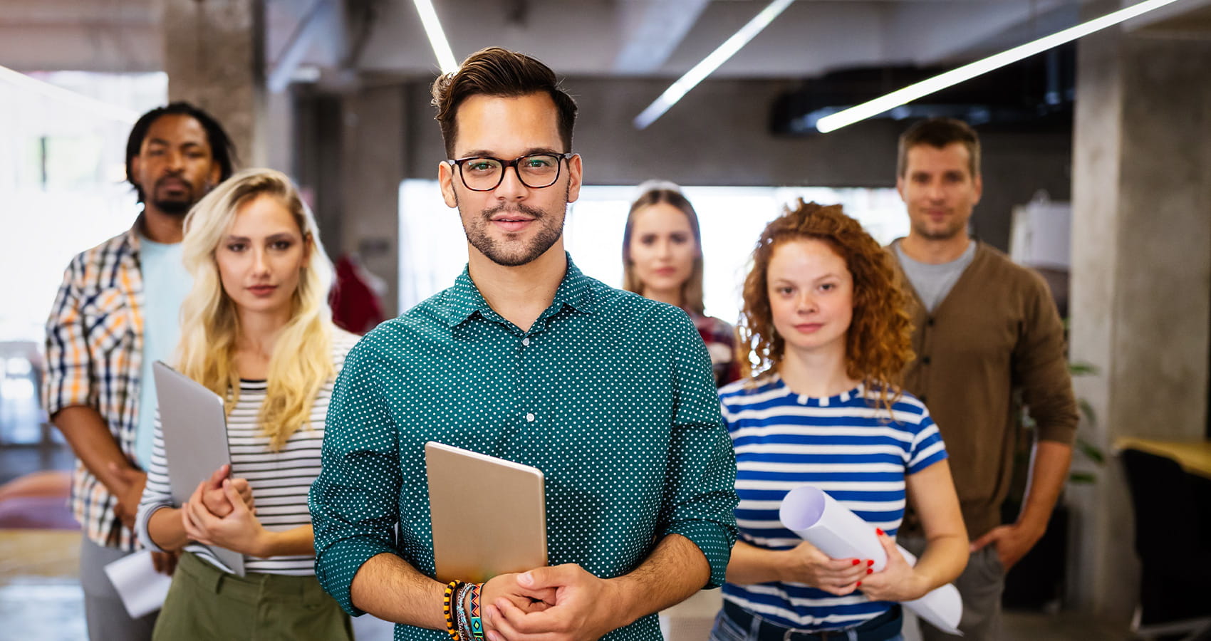 A diverse group of six young professionals, led by a man in a teal shirt, look confidently at the camera.