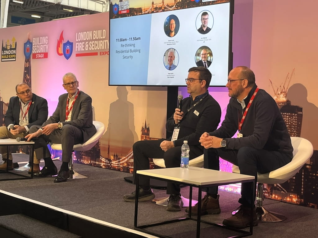 Panel discussion with four speakers seated on stage, presentation screen behind showing speaker details.