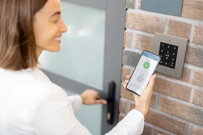 Person unlocking a door using a smartphone with a mobile access app near a wall-mounted keypad reader.