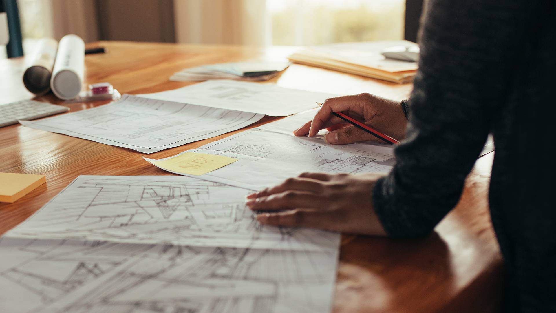 Personne examinant des plans architecturaux et des croquis sur un bureau en bois avec des plans enroulés à proximité