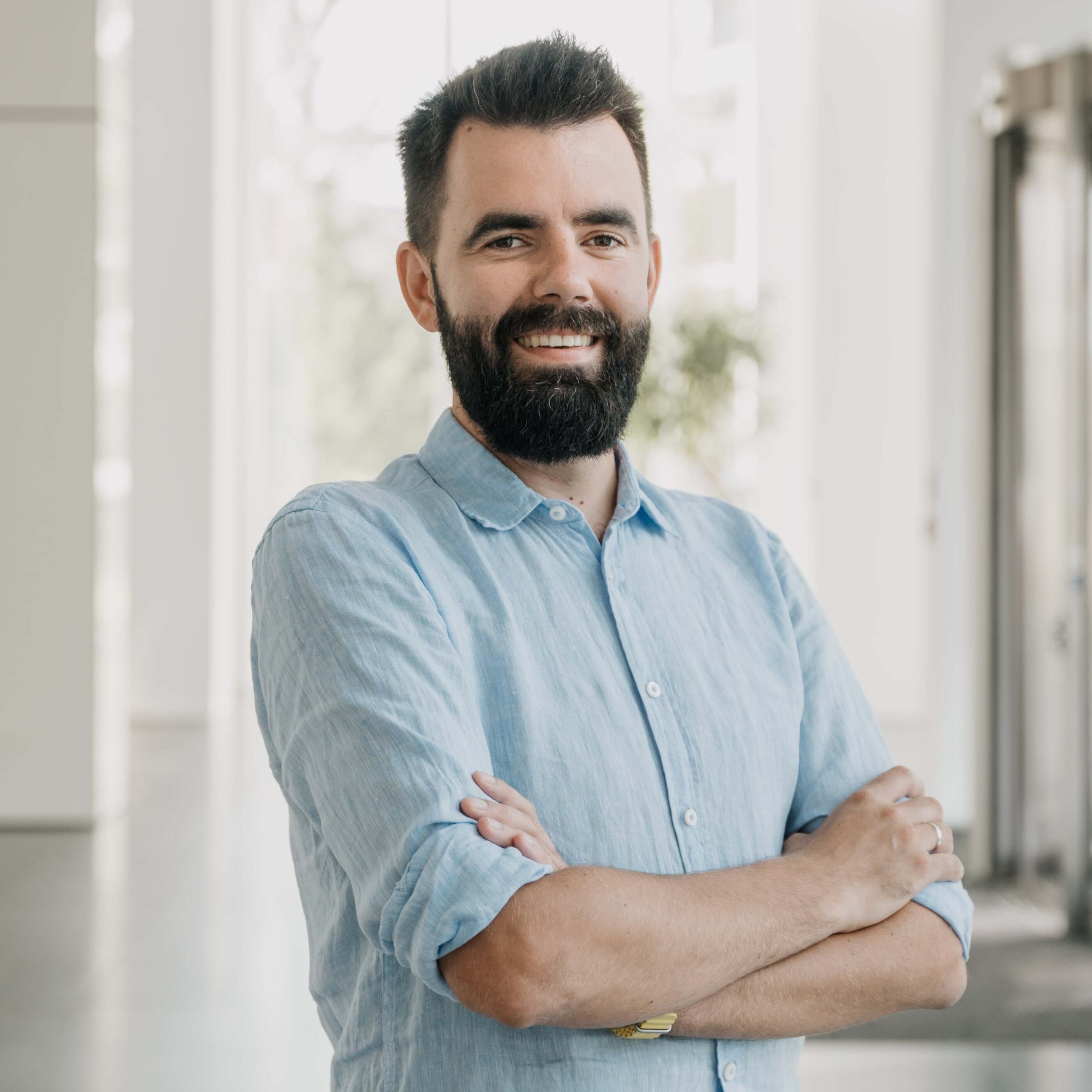 Personne portant une chemise boutonnée bleu clair avec les manches retroussées, se tenant les bras croisés dans un intérieur moderne et lumineux.