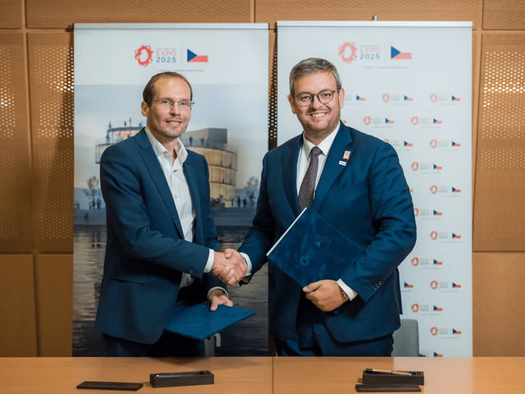 Dva jednotlivci ve formálních oblecích si podávají ruce na podpisové ceremonii s českými tematickými bannery za nimi.