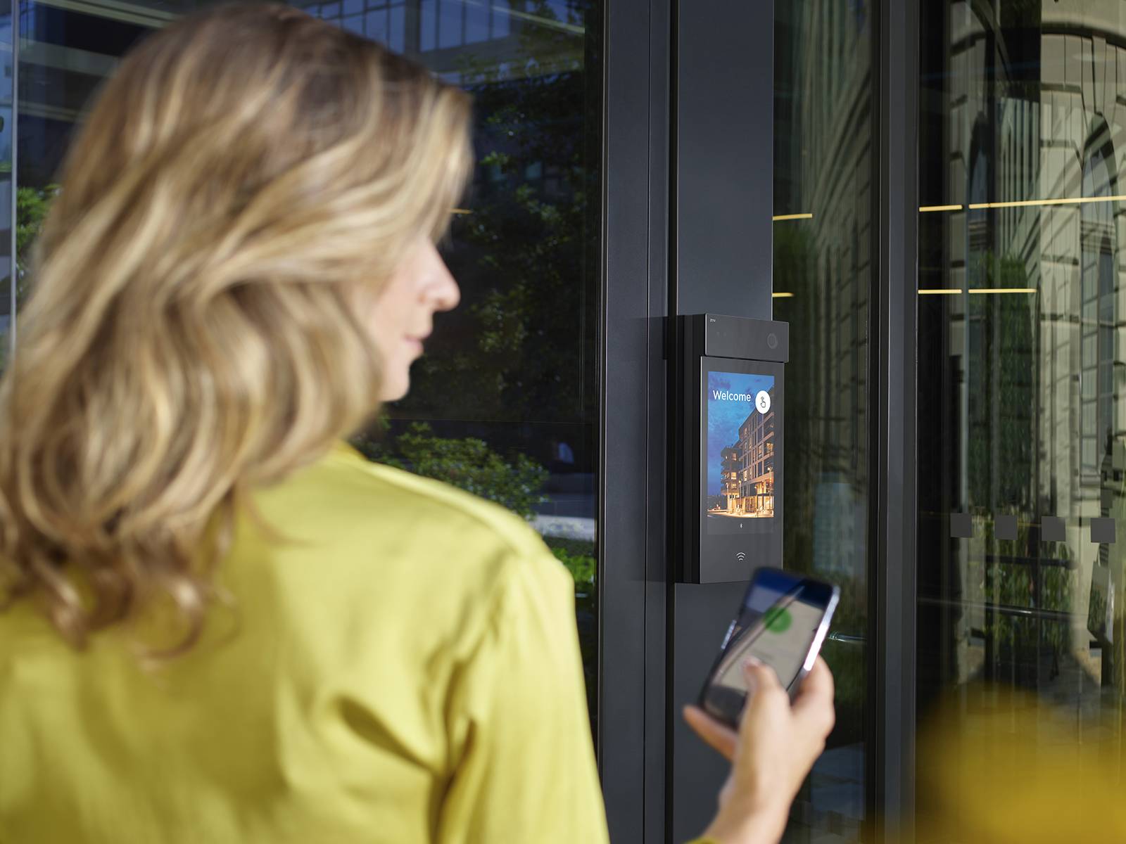 Osoba trzymająca smartfon w pobliżu szklanych drzwi z zamontowanym obok nich inteligentnym czytnikiem kontroli dostępu.