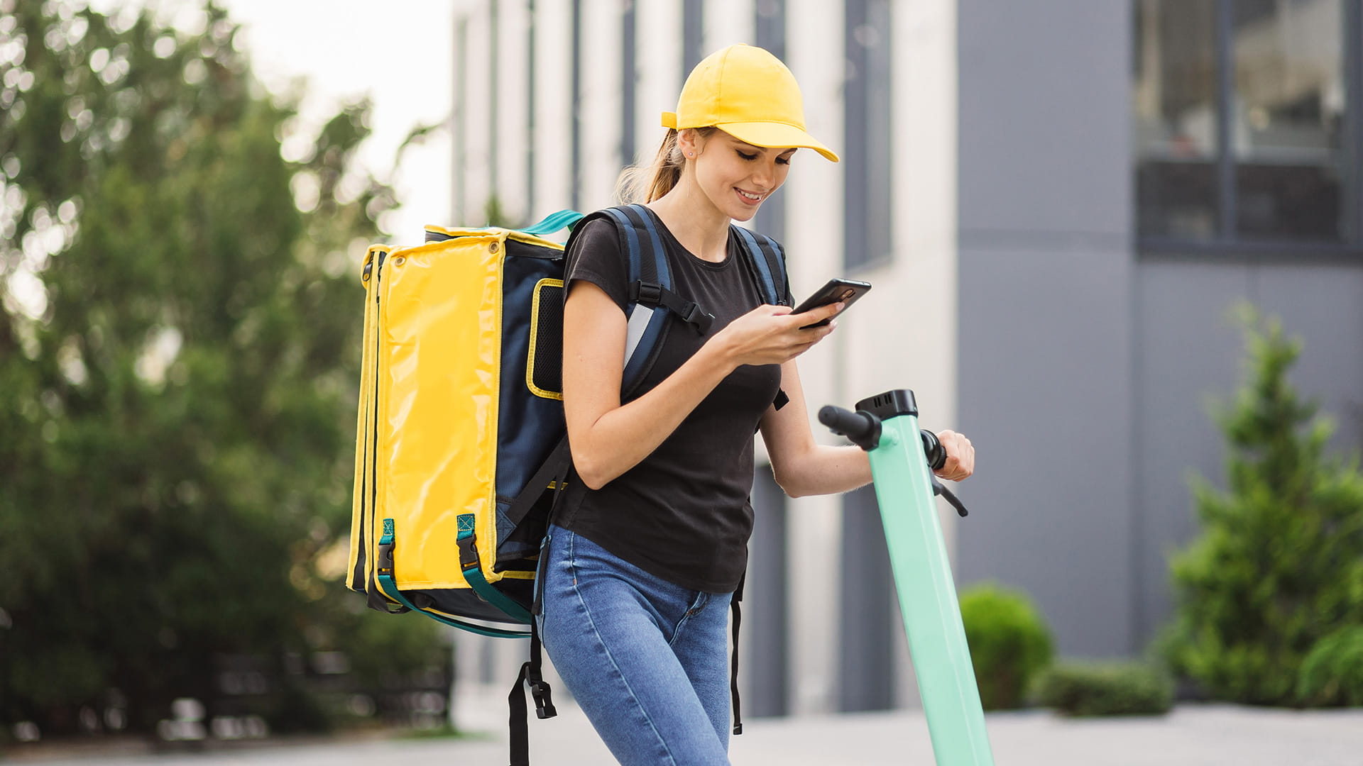 Lavoratore della consegna con uno zaino giallo che controlla uno smartphone mentre tiene uno scooter elettrico.