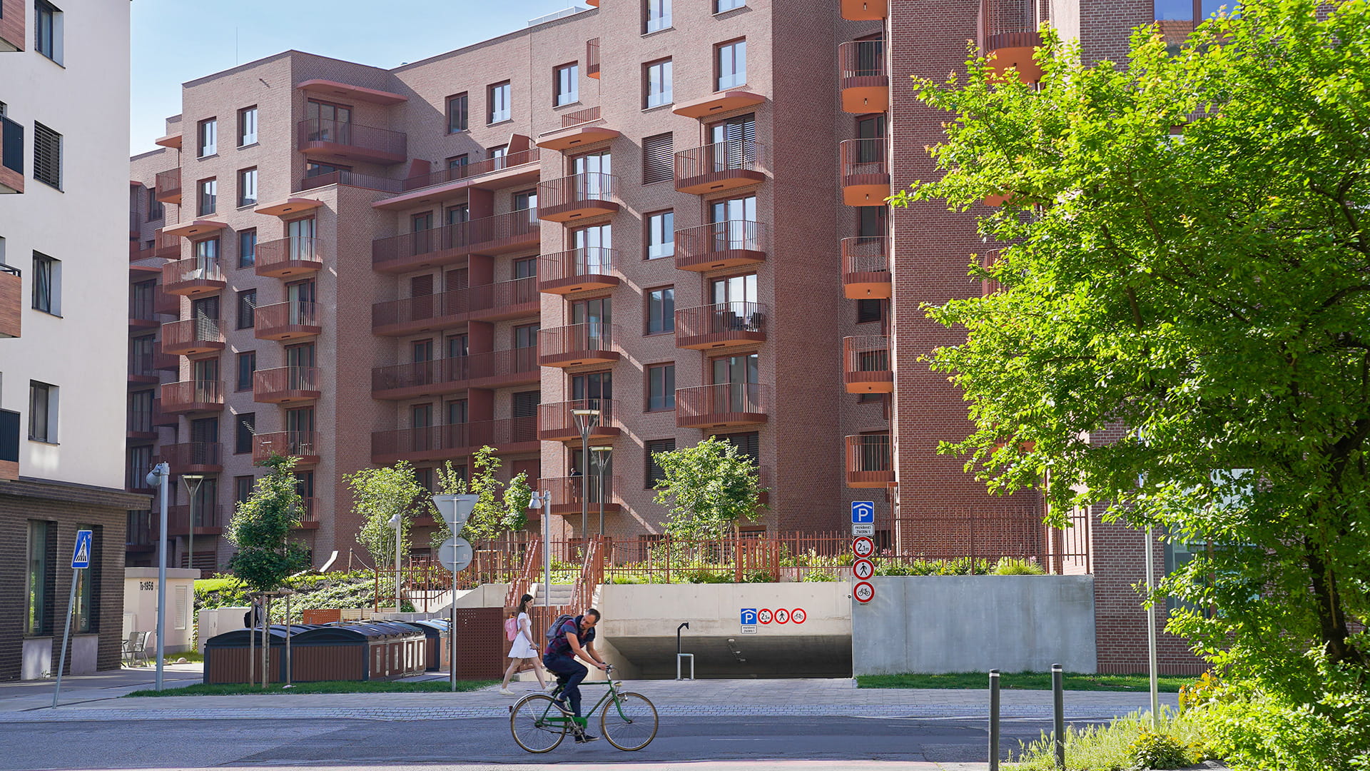 Moderner Wohnkomplex mit braunen Balkonen, viel Grün und einem Radfahrer im Vordergrund.