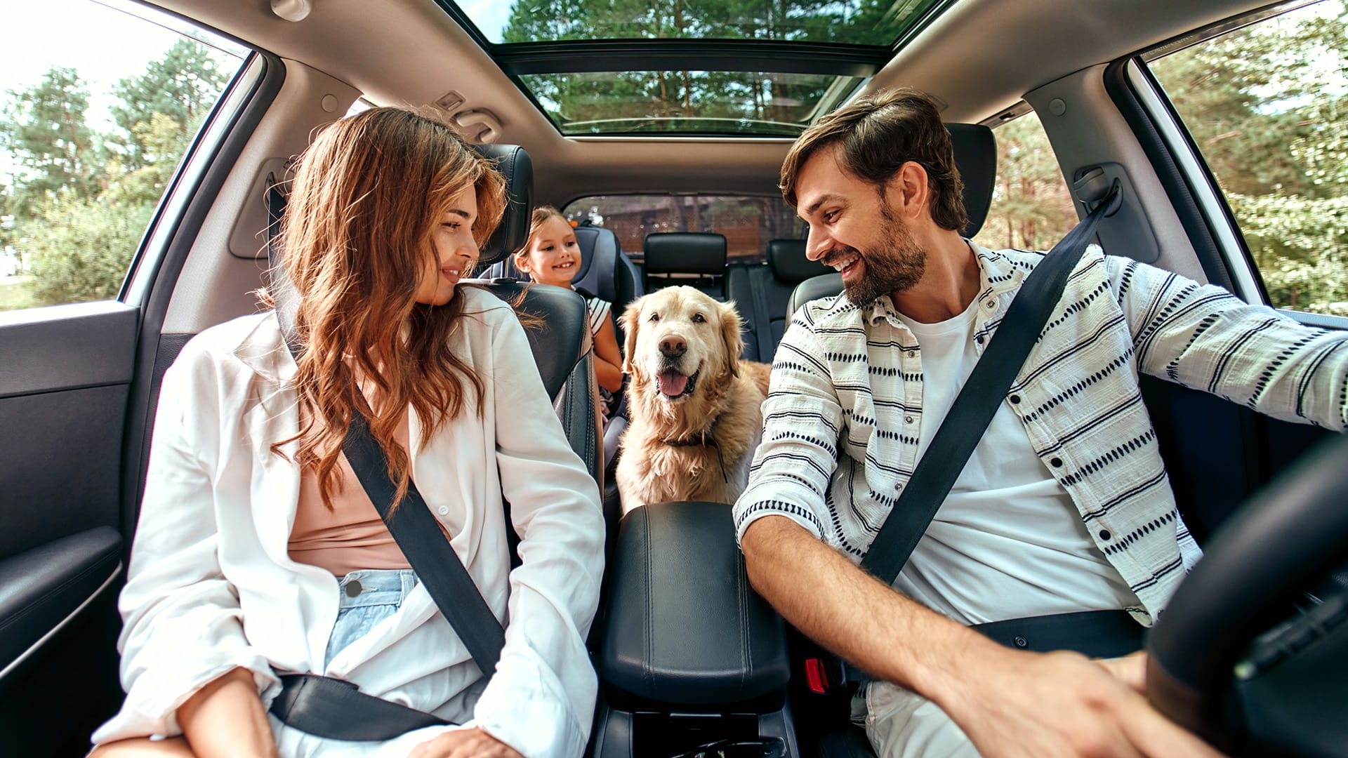 家族が車に座っていて、中央の座席に犬がいて、後ろに子供がいて、森の中を運転している。