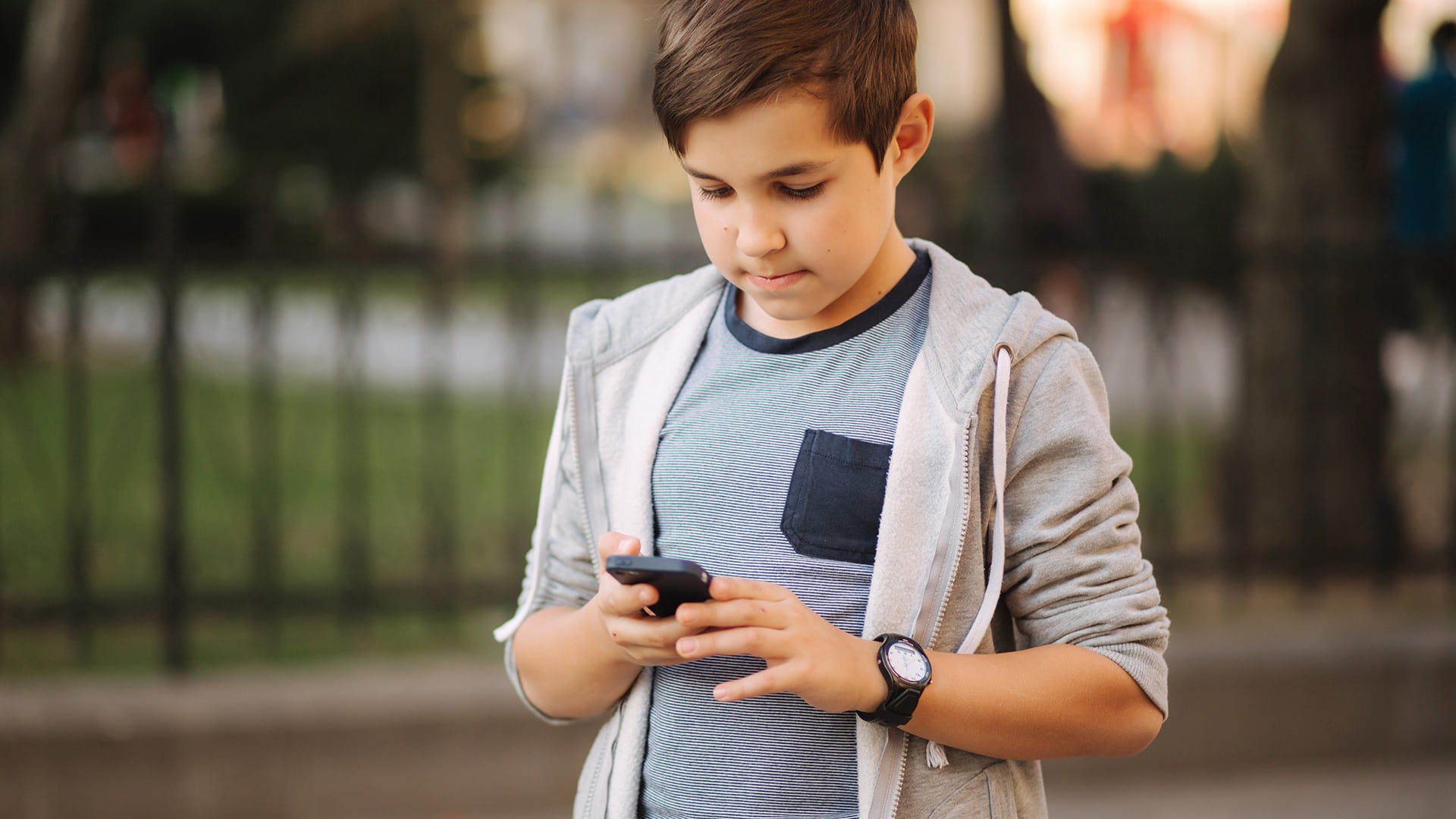 子供が屋外に立ってカジュアルなグレーのフーディを着てスマートフォンを使っています。