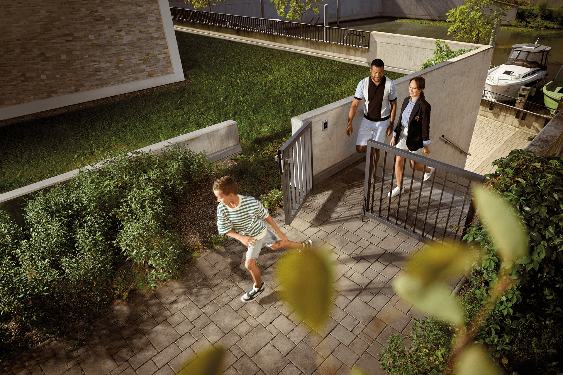 Drie mensen die een moderne omheinde woning binnenkomen met een geplaveide loopbrug en groen rondom de ingang.