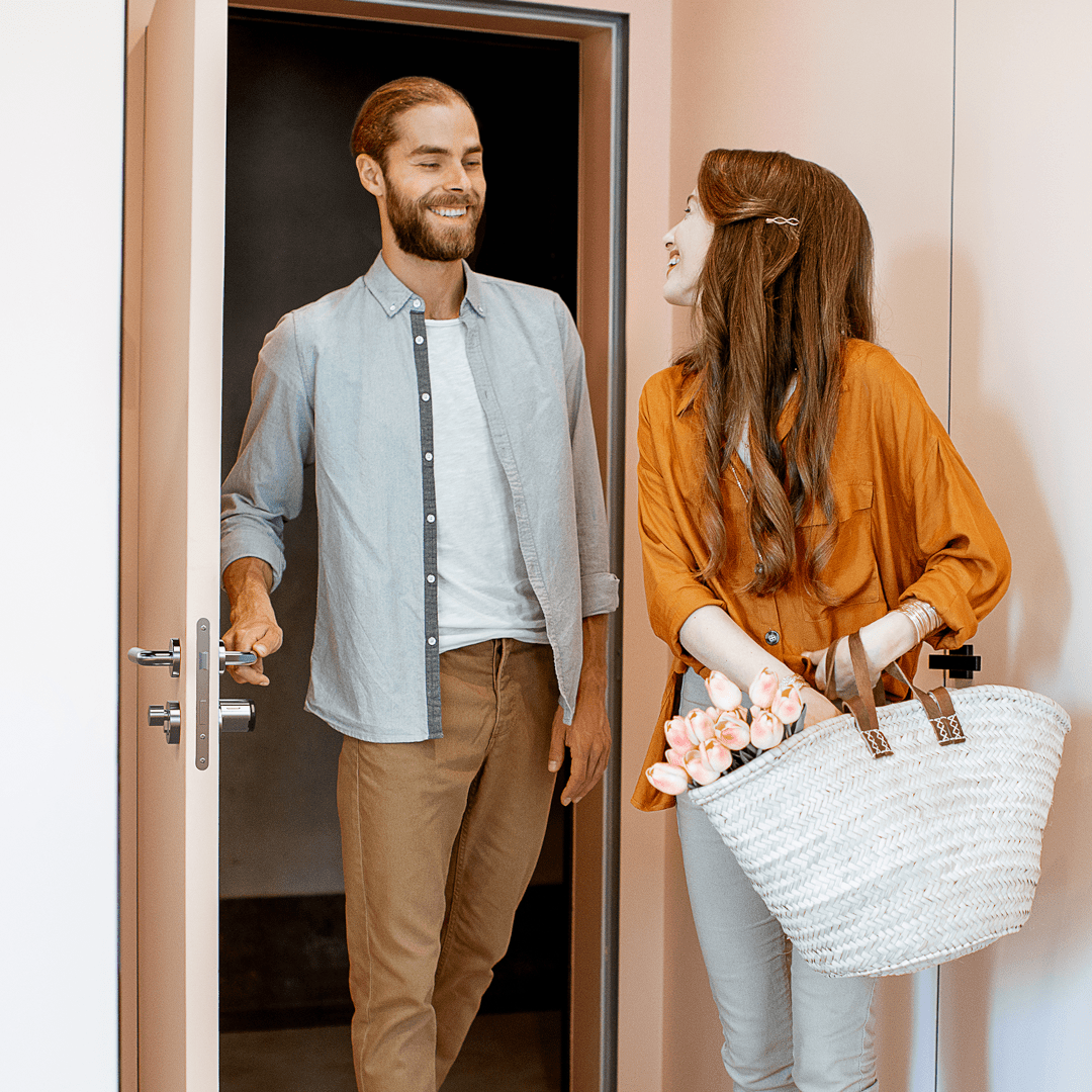 Koppel dat een appartement binnenkomt, man die de deur opent voor een gast met een tas en bloemen