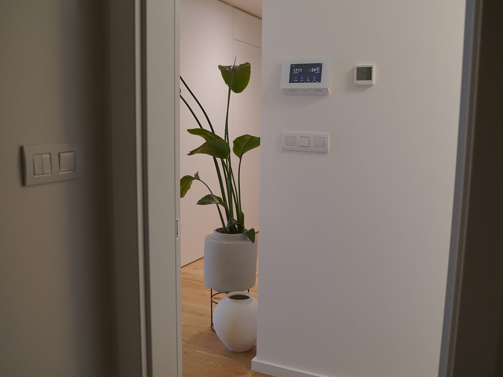 Corredor interno com paredes brancas, um termostato inteligente e painel de controlo instalados, e uma planta decorativa num vaso branco.