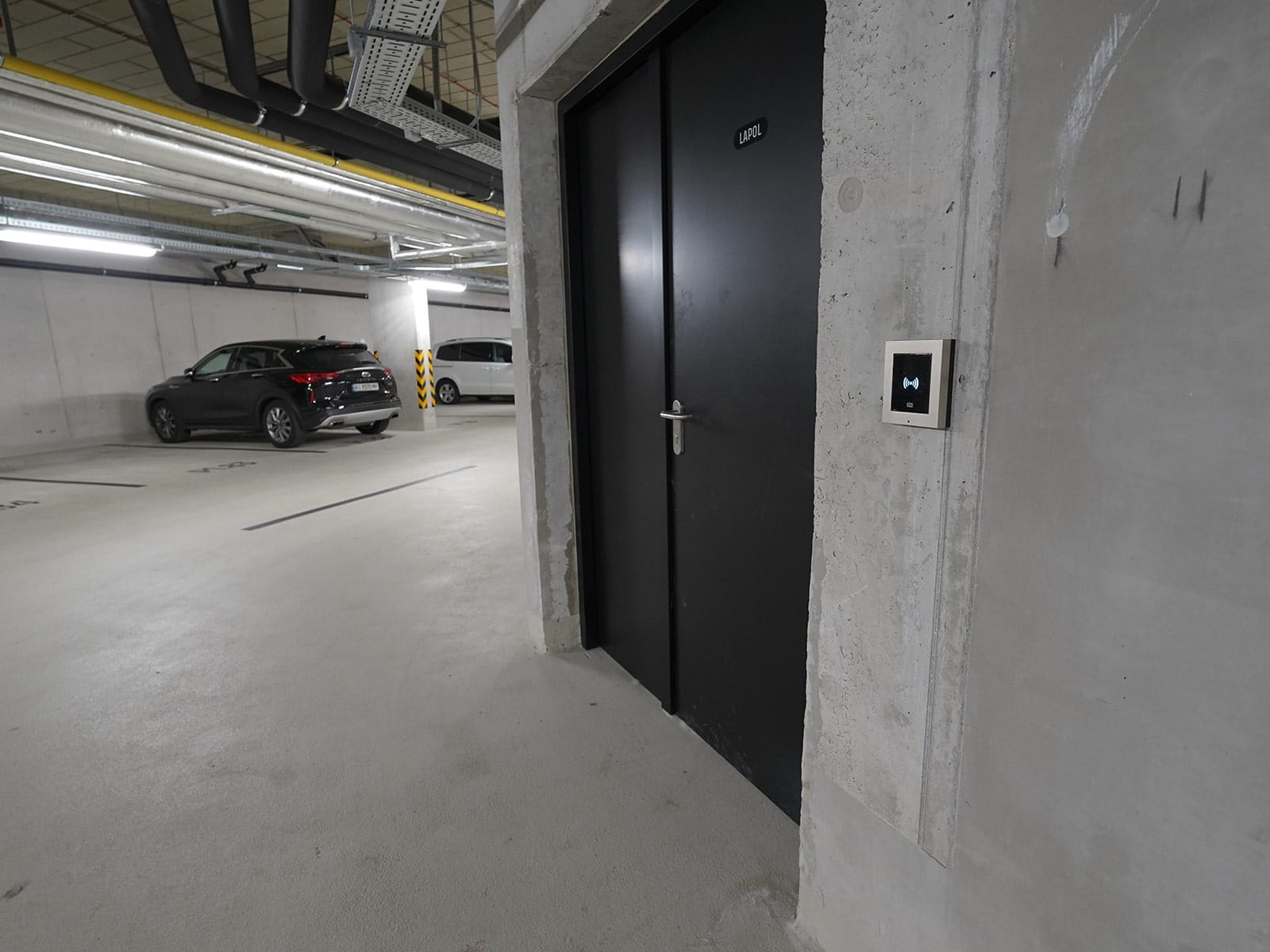 Garagem subterrânea com carros pretos e brancos e uma porta preta com um painel de acesso eletrónico.