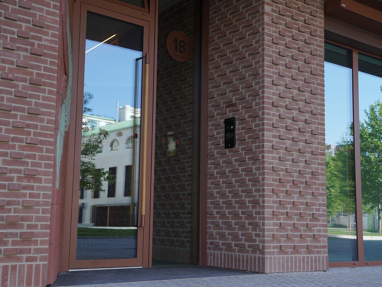 Entrada moderna do edifício com portas de vidro e fachada de tijolos com um painel de acesso eletrónico.