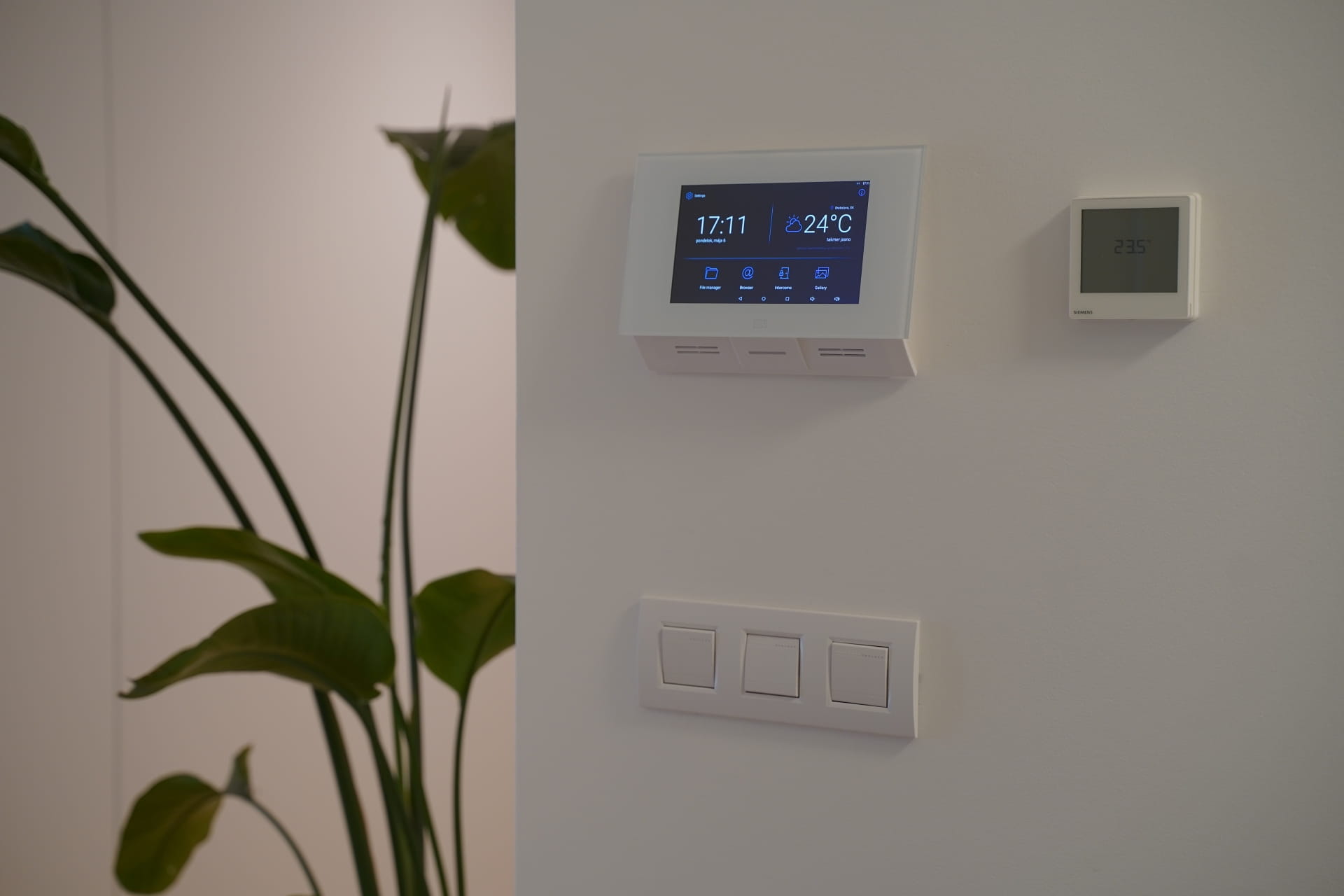 Parede branca com um painel de controlo doméstico inteligente, termóstato e interruptores de luz ao lado de uma planta verde.