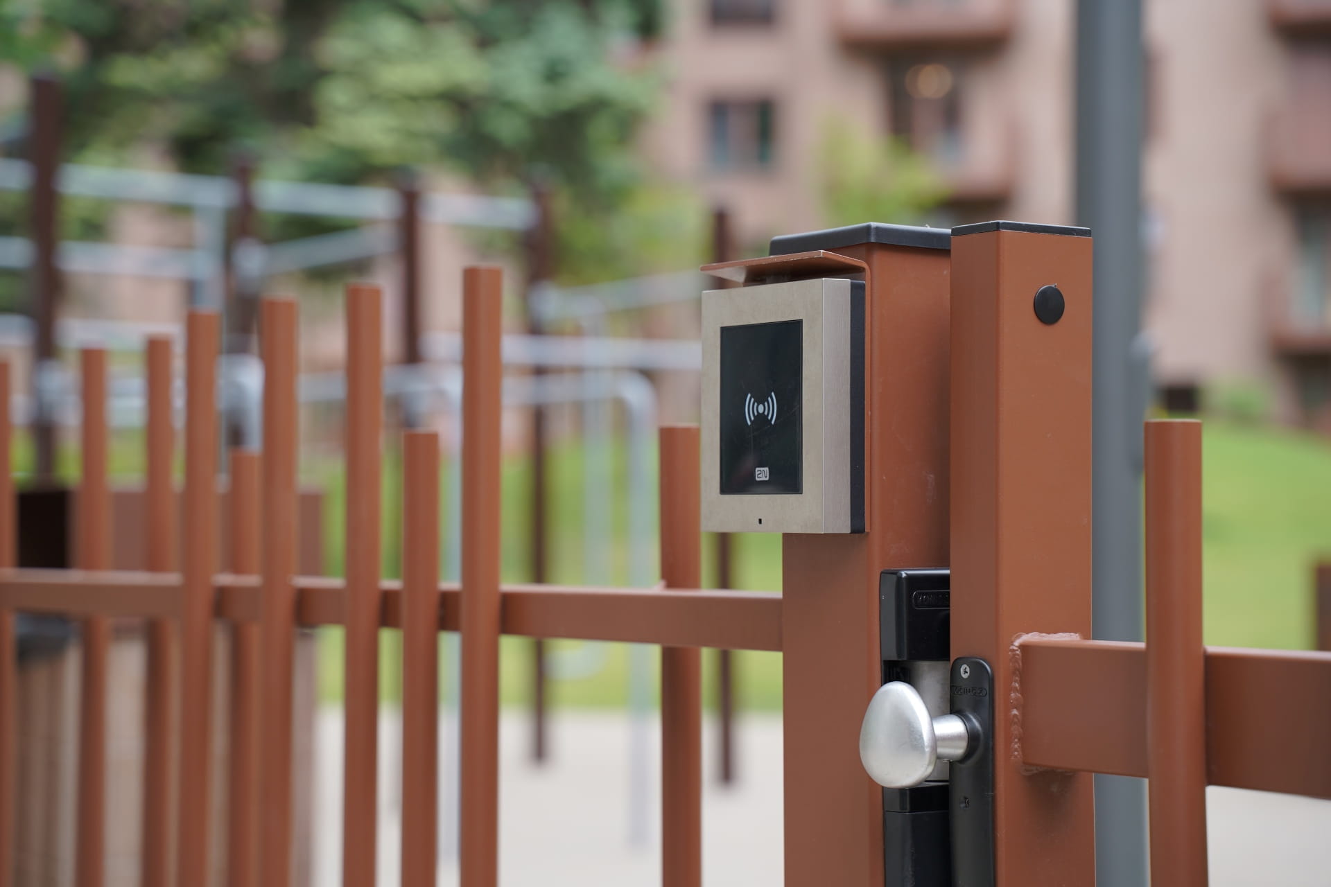 Leitor de controlo de acesso montado numa porta de metal castanho numa área exterior com edifícios ao fundo.
