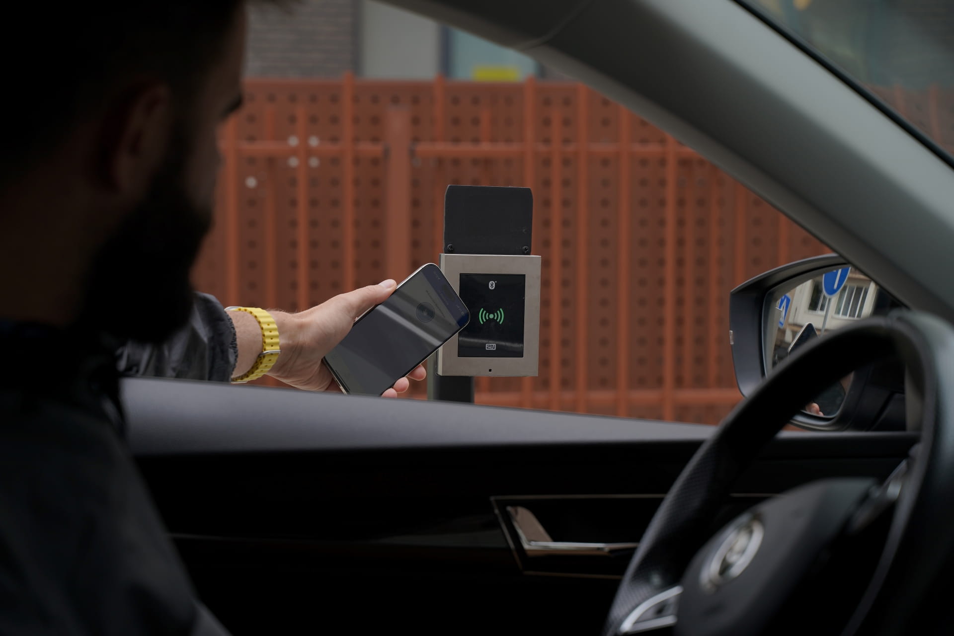 Uma pessoa dentro de um carro a segurar um smartphone perto de um leitor de controlo de acesso ao ar livre num portão.