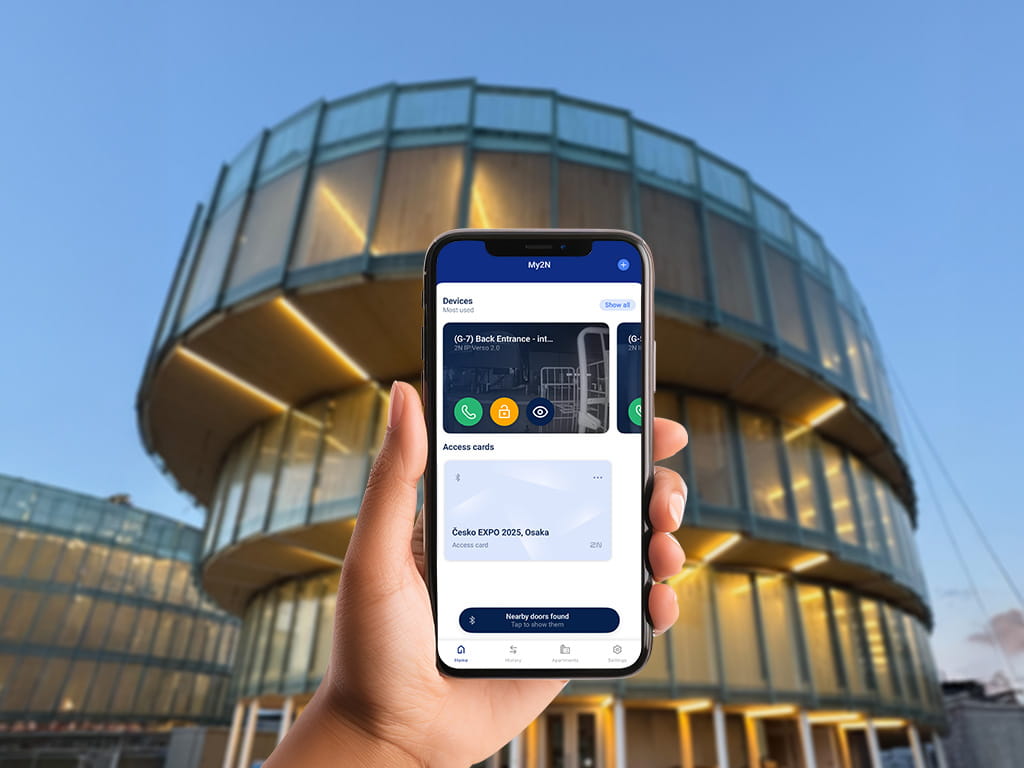 Ręka trzymająca smartfona z aplikacją do kontroli dostępu wyświetlaną przed nowoczesnym okrągłym szklanym budynkiem o zmierzchu.