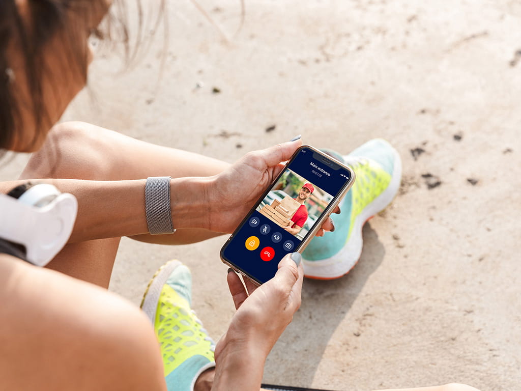 Osoba trzymająca smartfon z interfejsem wideorozmowy z kolorowymi przyciskami, siedząca na świeżym powietrzu w stroju sportowym.