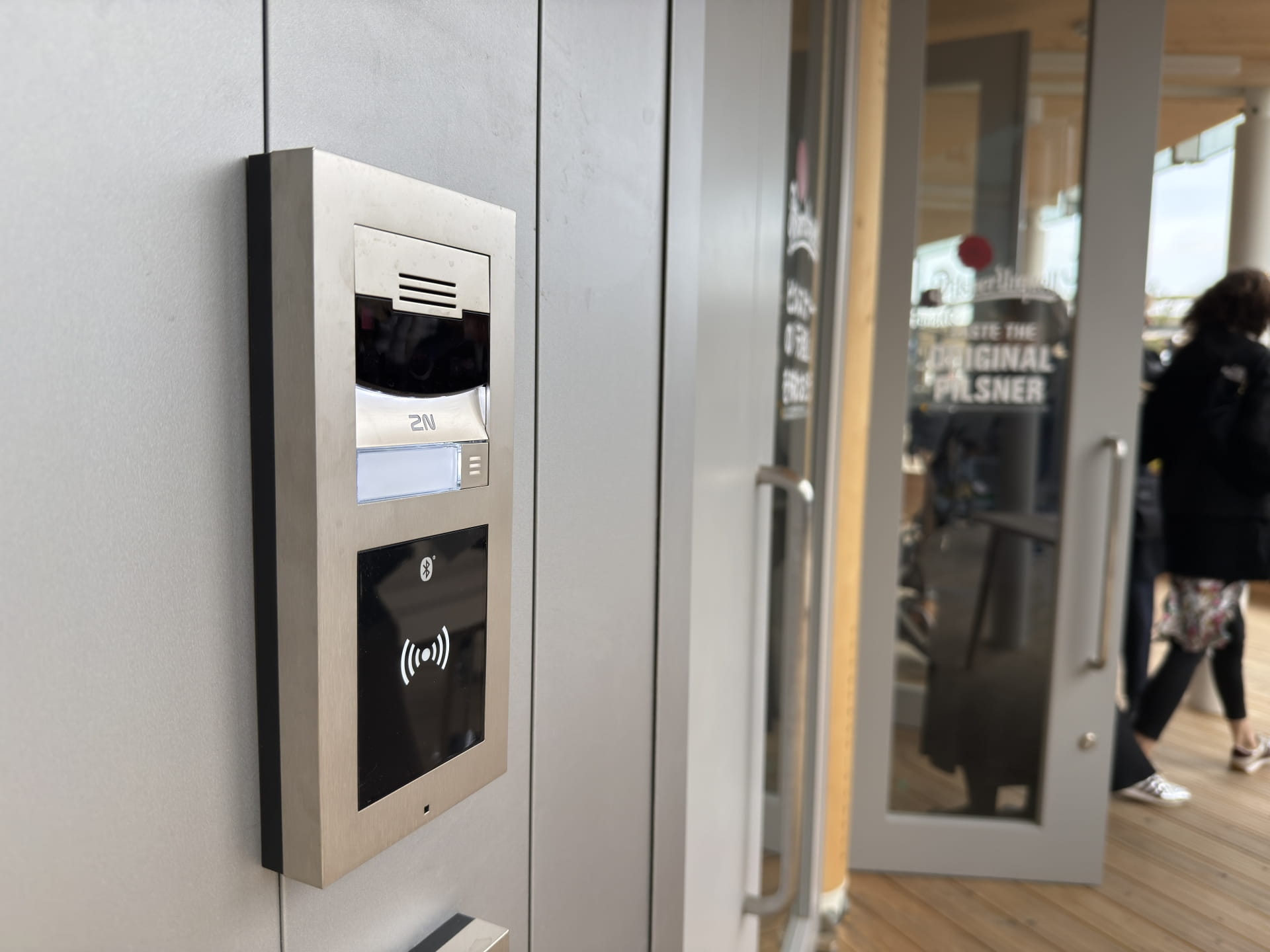 Zbliżenie na system domofonowy ze stali nierdzewnej zamontowany na szarej ścianie obok szklanych drzwi wejściowych.
