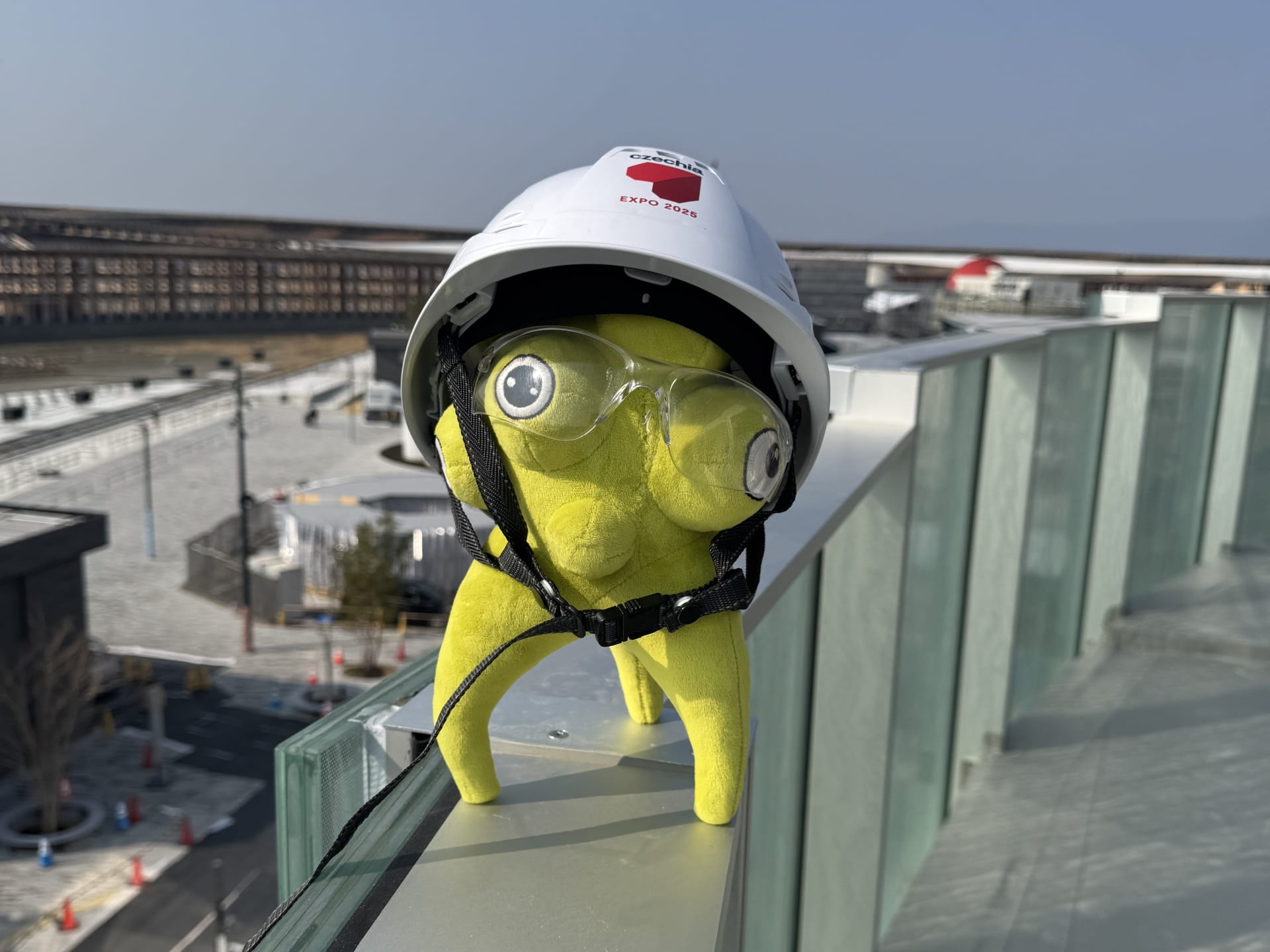 Żółta pluszowa zabawka nosząca białą kask i gogle umieszczona na szklanej balustradzie na placu budowy.