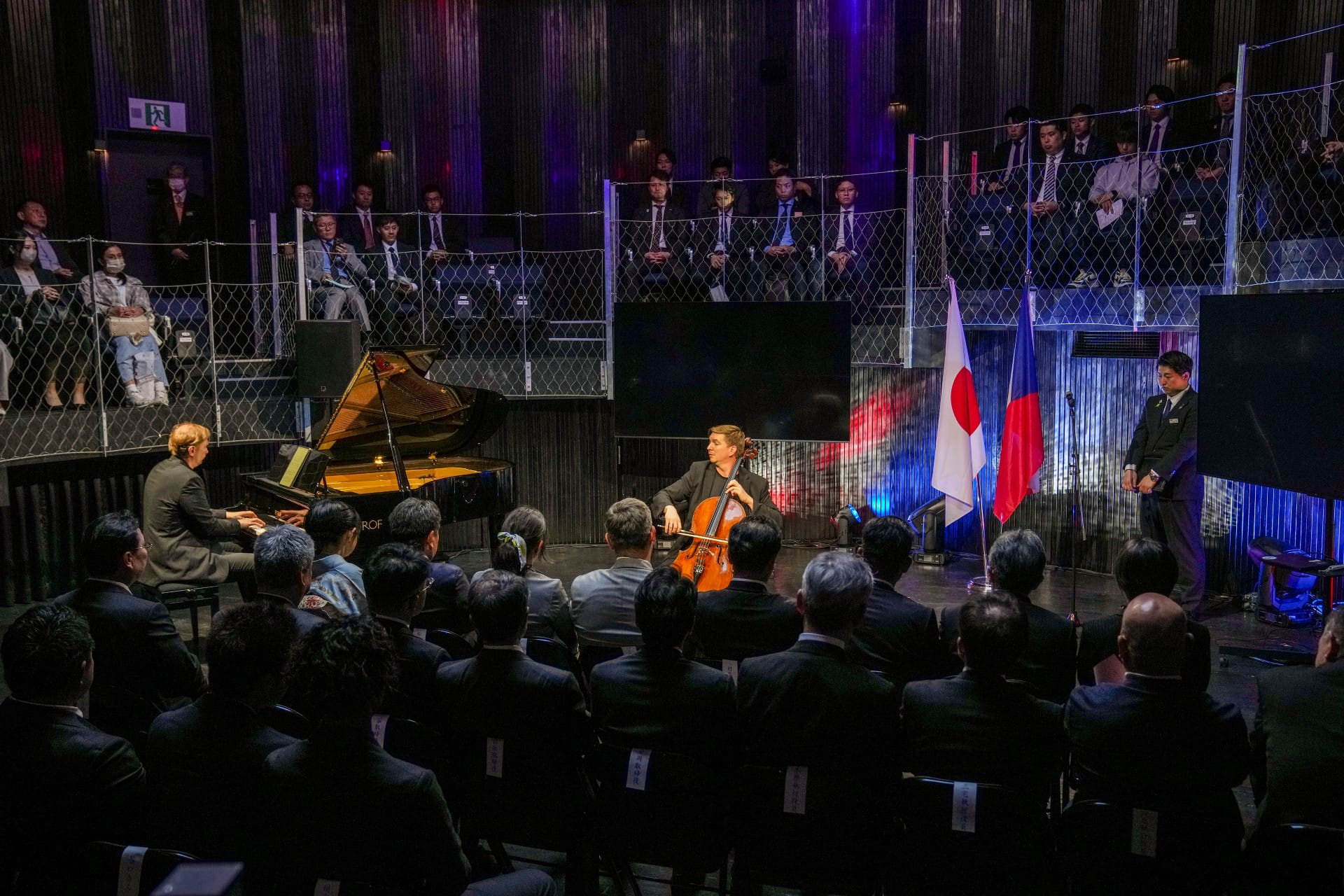  Impreza w pomieszczeniu, podczas której siedząca publiczność ogląda występ na żywo pianisty i wiolonczelisty, a na scenie wywieszone są flagi.