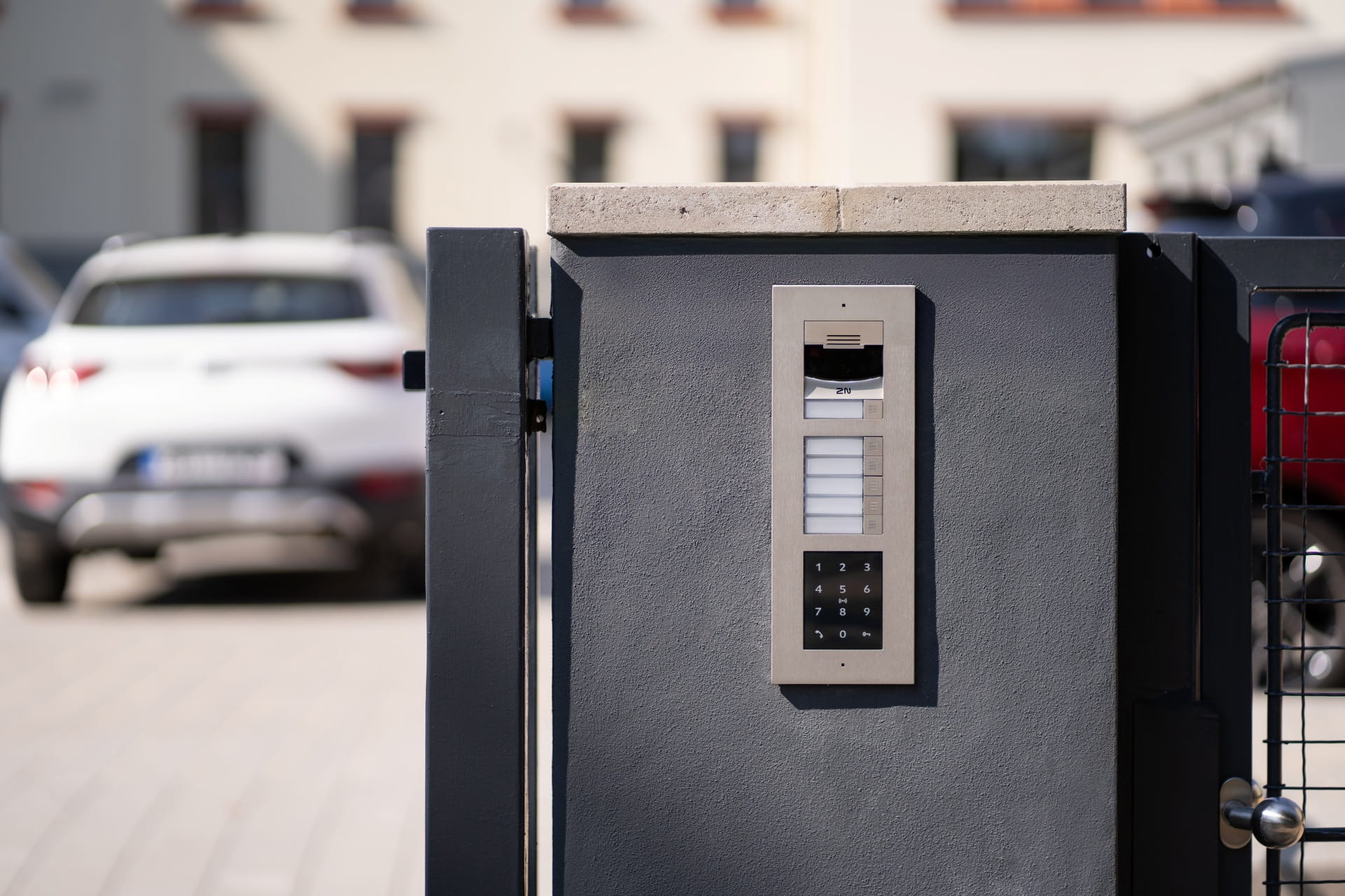 Portail extérieur avec un interphone et un système d'accès par clavier, voitures garées en arrière-plan.