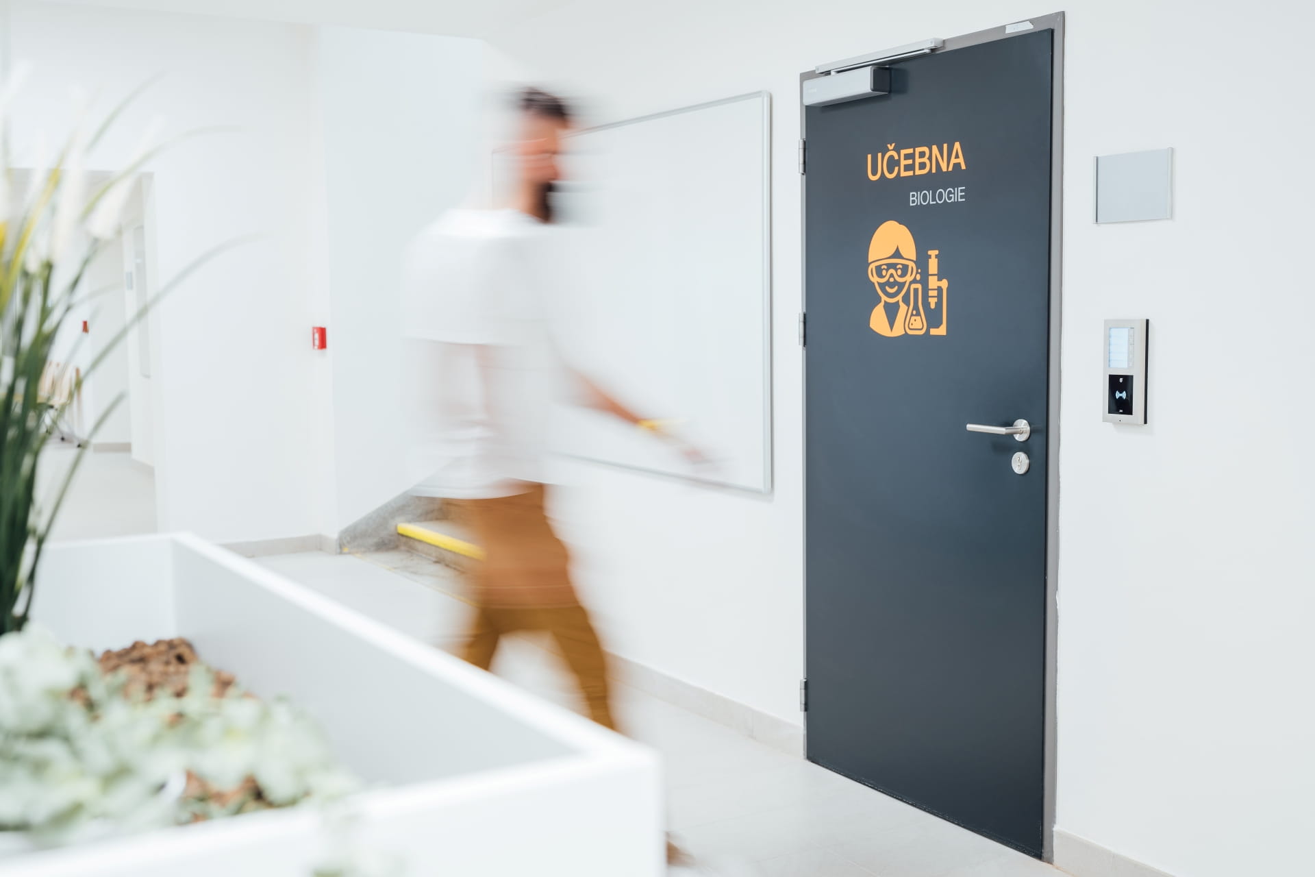 Une personne passant devant une porte de salle de classe étiquetée "UČEBNA BIOLOGIE" avec un lecteur de contrôle d'accès sur le mur.