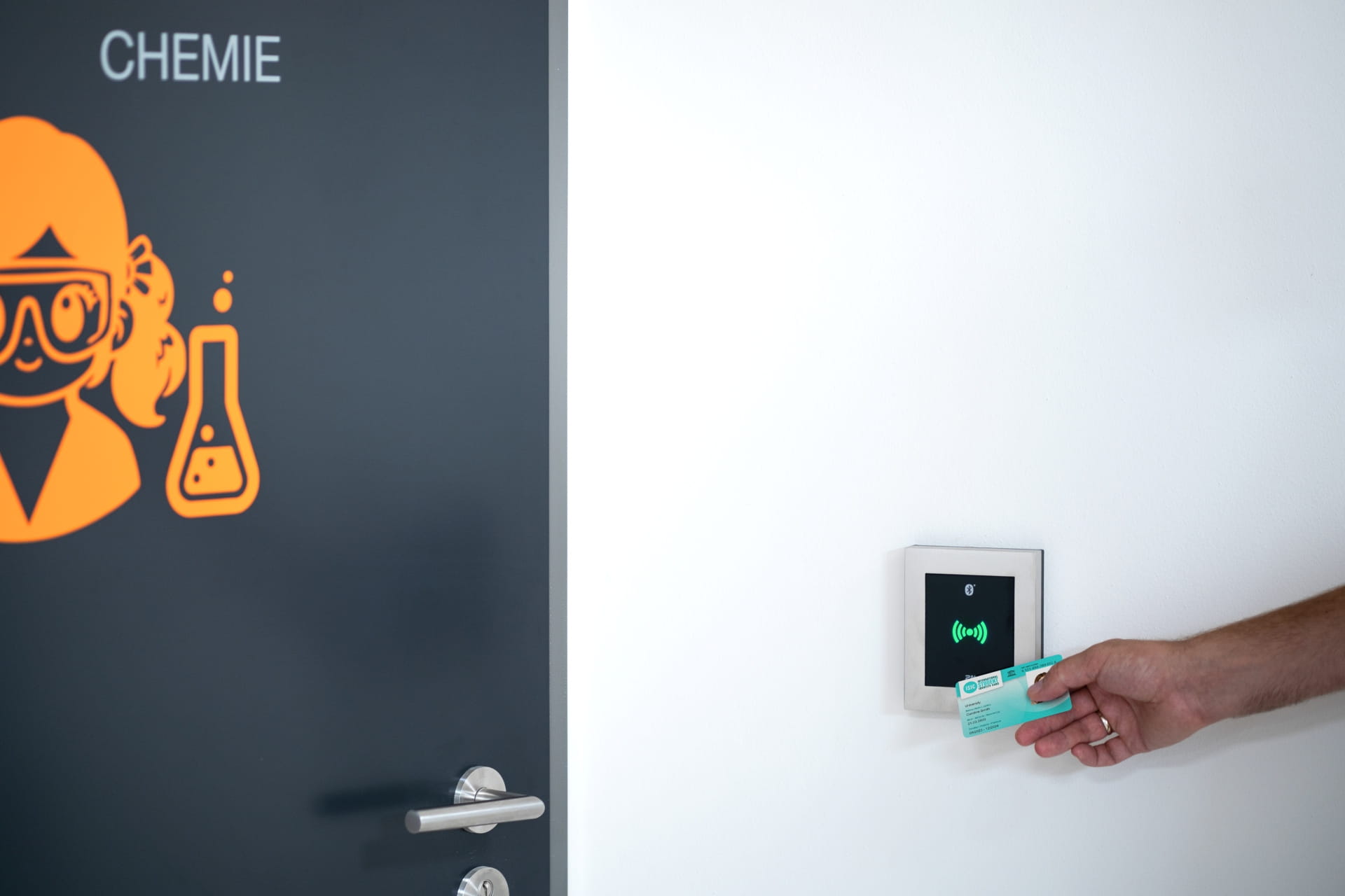 Main tenant une carte près d'un lecteur mural pour le contrôle d'accès à côté d'une porte étiquetée "CHEMIE."