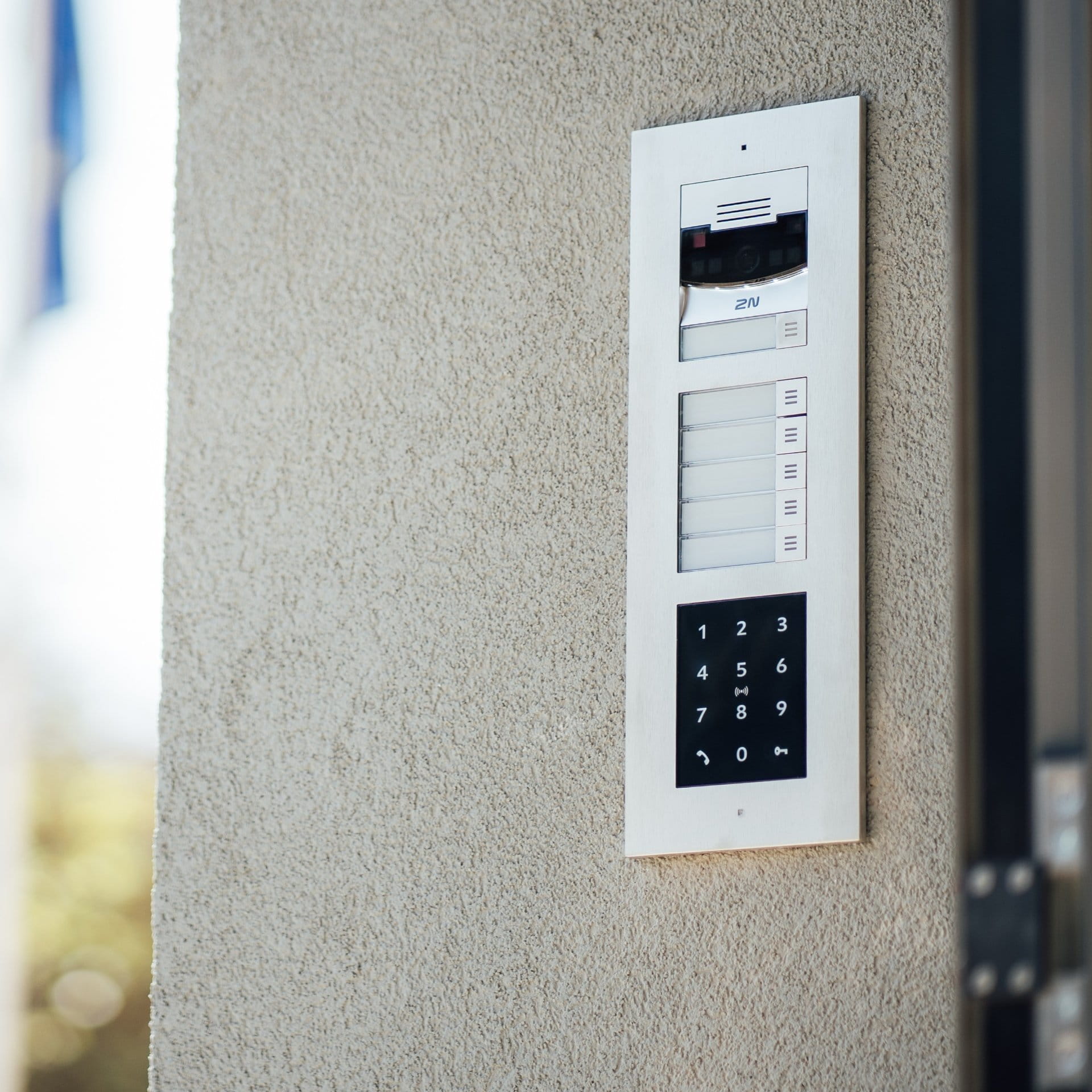 Interphone mural avec caméra, plaques nominatives et clavier numérique sur un mur extérieur texturé.